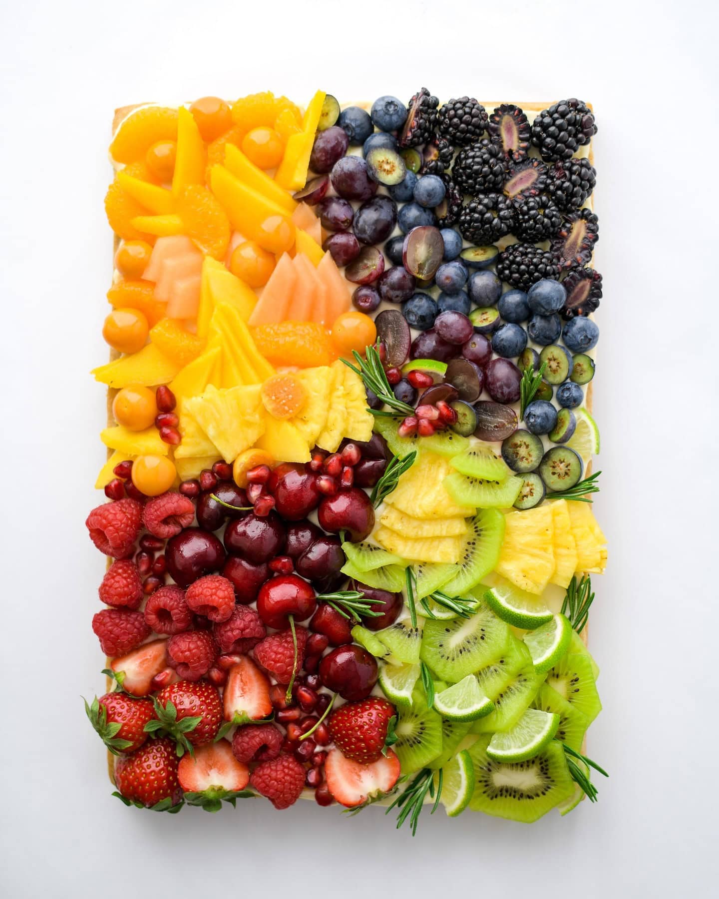 A vibrant fruit platter for a rainbow baby shower, with assorted fruits neatly arranged in sections. From top left clockwise: orange slices and cherry tomatoes, blueberries and blackberries, kiwi slices and pineapple chunks, strawberries, raspberries, and cherries, with some sprigs of rosemary.