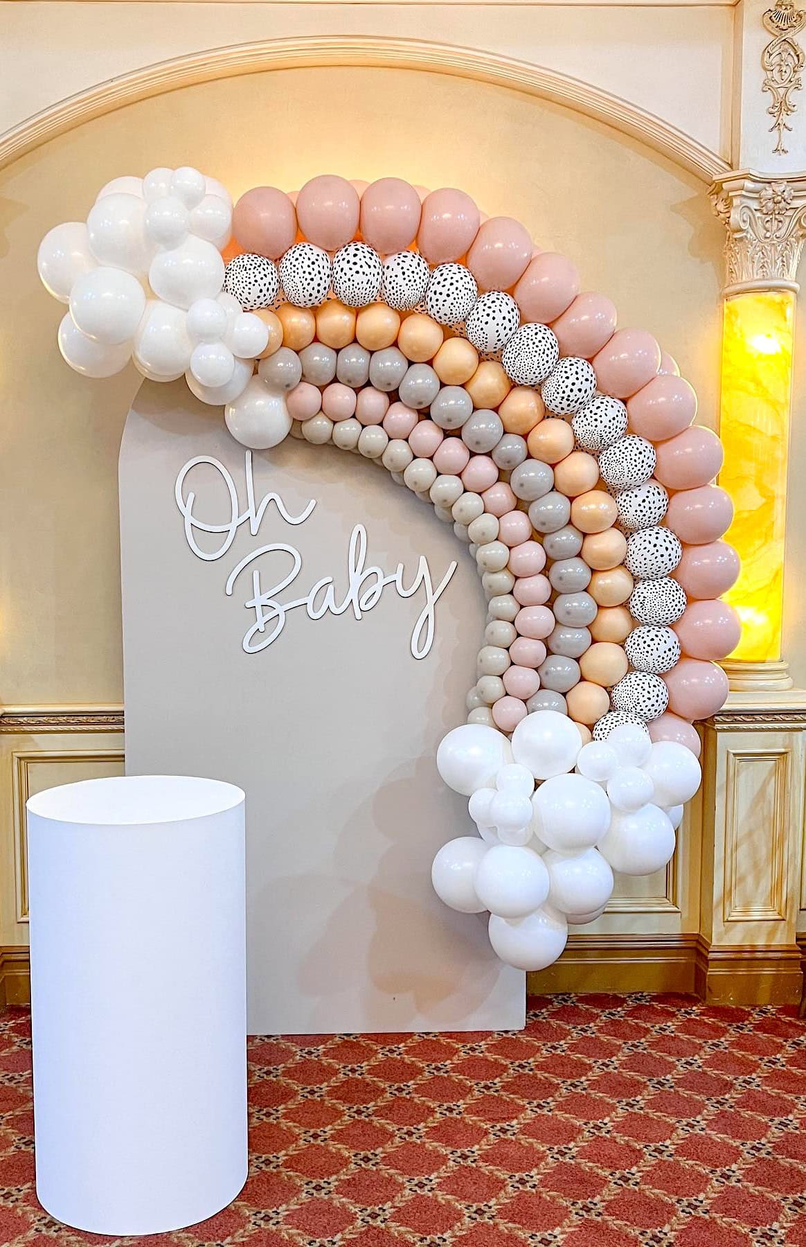 A stylish rainbow baby shower backdrop features a large arch of balloons in shades of white, pink, peach, and polka dots, forming a vibrant rainbow shape. The words "Oh Baby" are displayed on a beige board beside a white cylindrical table, set against an ornate wall.