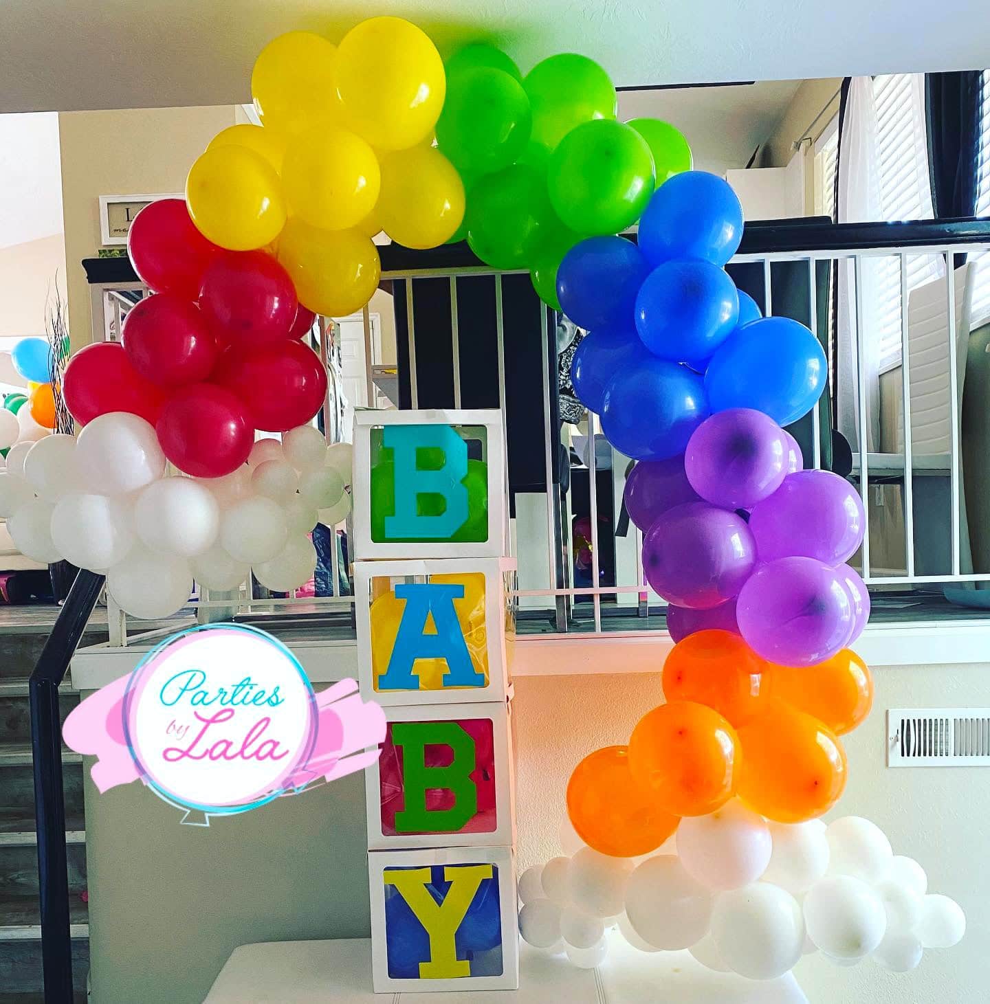 A vibrant balloon arch with red, white, green, blue, purple, and orange balloons frames a stack of four transparent boxes spelling "BABY" in green and yellow. Perfect for a rainbow baby shower, the sign reads "Parties by Lala" in pink and blue.