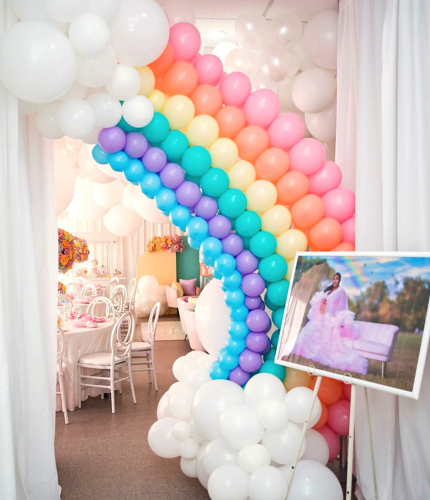 A room prepared for a rainbow baby shower features a colorful balloon arch arranged in a vibrant rainbow pattern with white clouds. Under the arch is a table set with white chairs and floral centerpieces. A framed photo of the person in formal attire is displayed on the right.