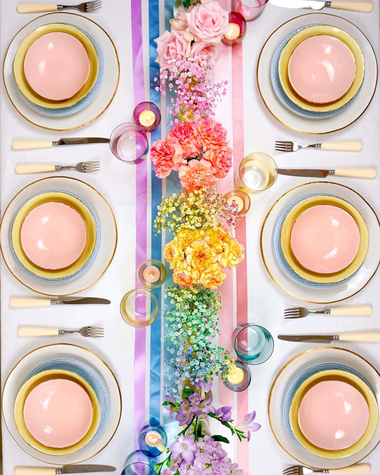 A vibrant table setting for a rainbow baby shower features six place settings with pink plates, yellow bowls, blue saucers, and white-handled utensils. The table is adorned with colorful flowers, candles, and pastel ribbons running down the center, creating a cheerful and elegant atmosphere.