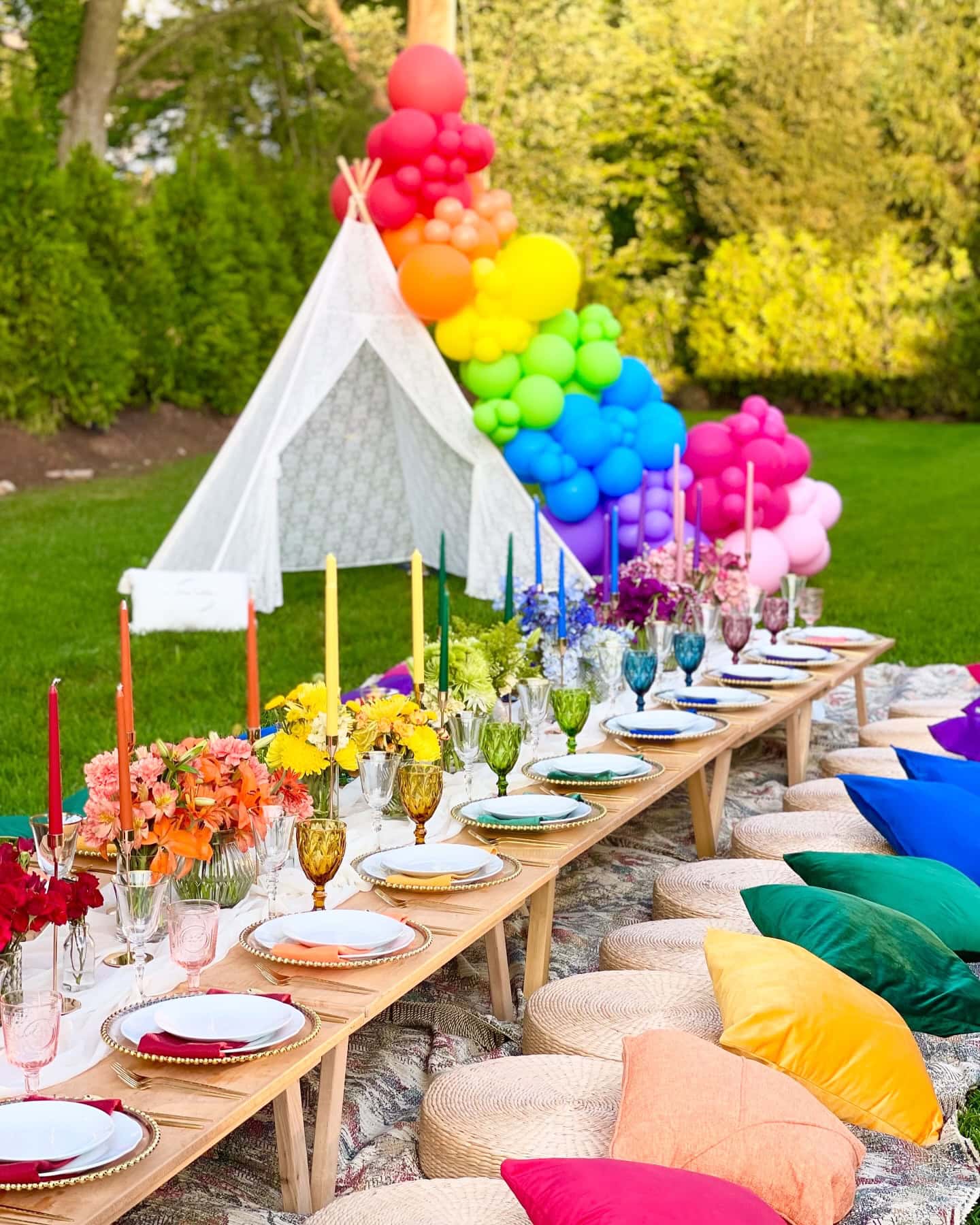 A long outdoor picnic table set for a rainbow baby shower features colorful candles, flowers, and tableware. Cushions and blankets for seating add a cozy touch. A white teepee adorned with rainbow balloons is visible in the background, set against lush green foliage.