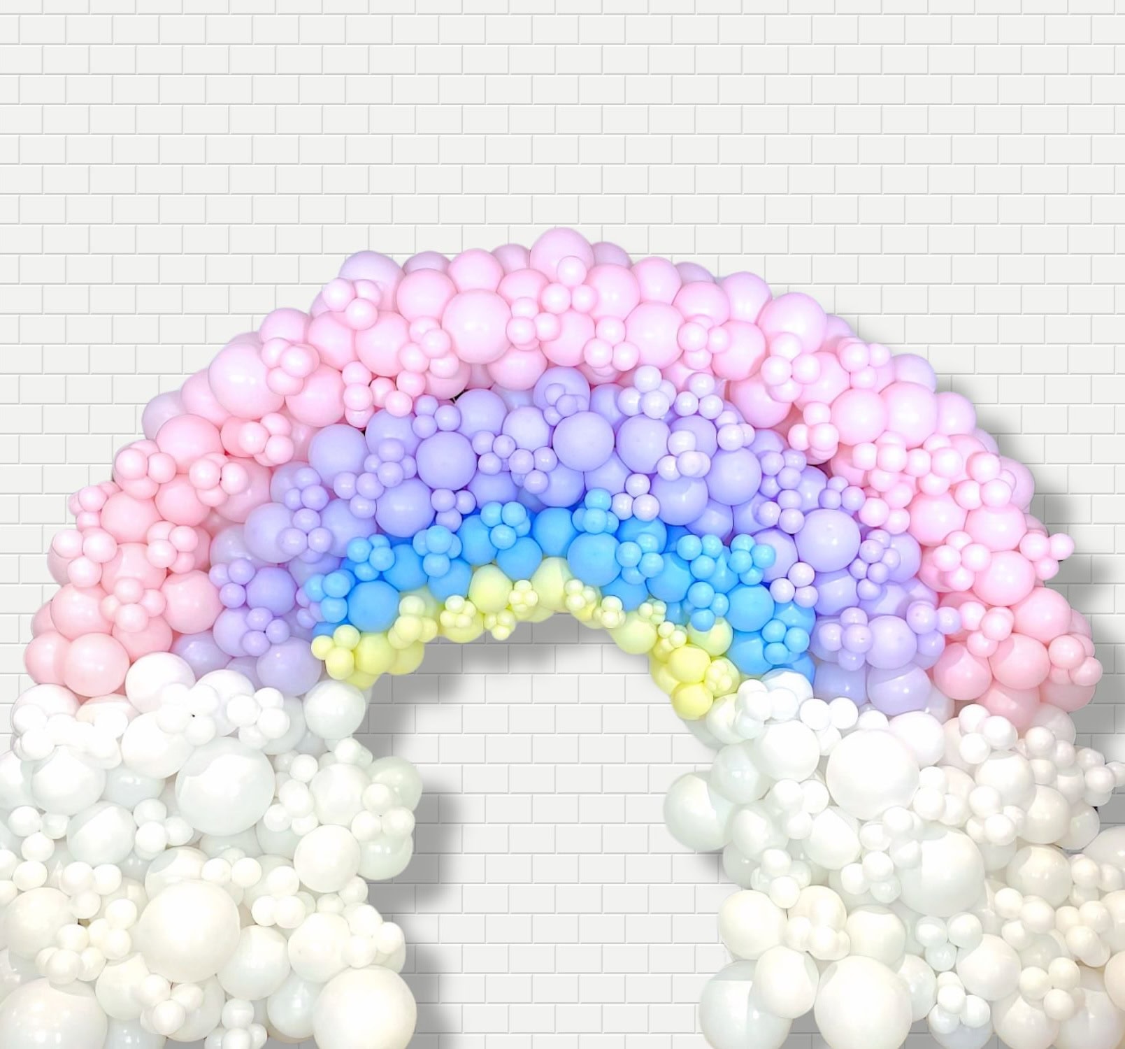 A balloon rainbow perfect for a rainbow baby shower, made of clusters of white, pink, lavender, blue, and yellow balloons. Arranged in an arch against a white brick background, the varying sizes create a whimsical and colorful display.
