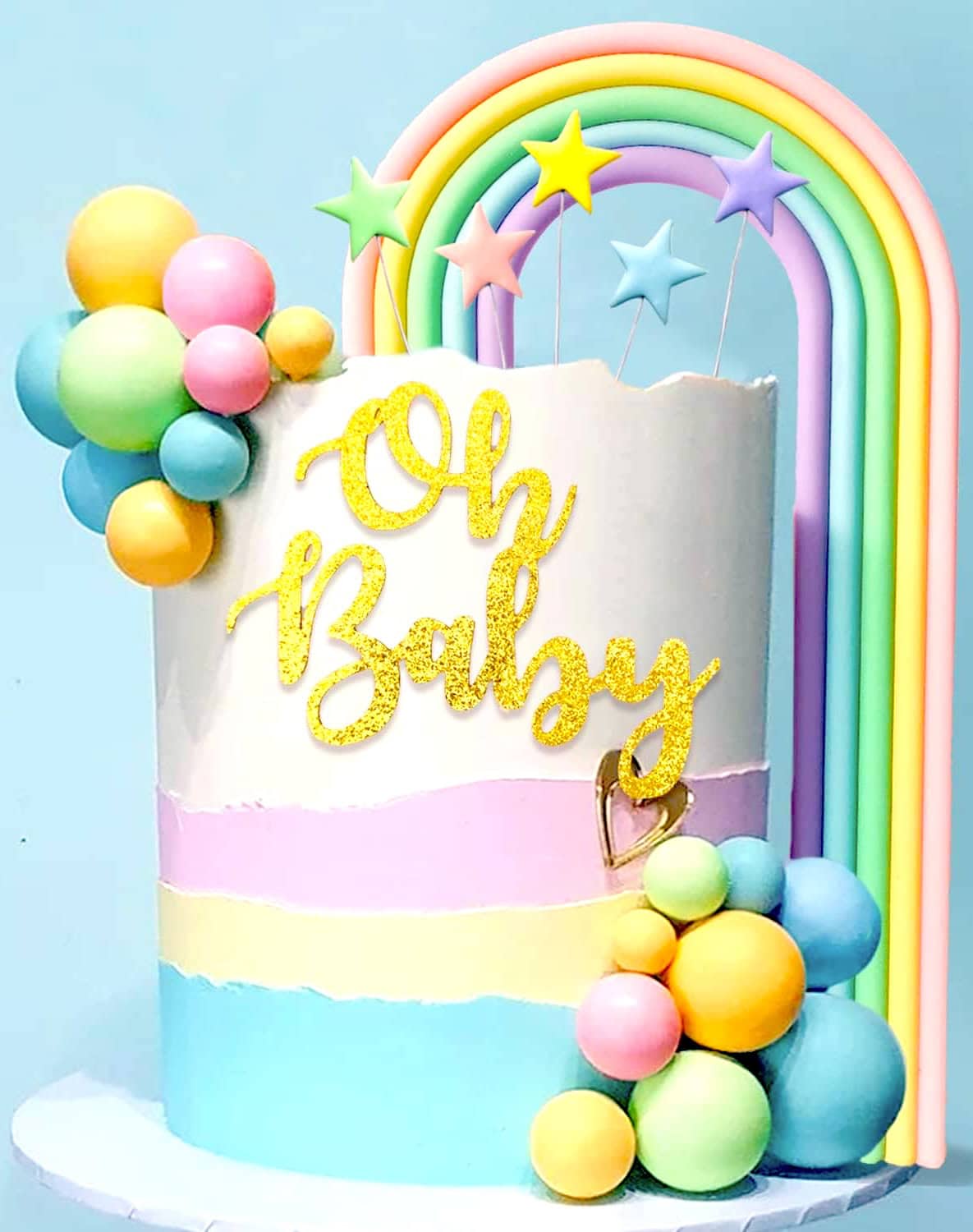 A pastel-colored rainbow baby shower cake features the words "Oh Baby" in gold glitter cursive on the side. Rainbow arches and colorful star toppers adorn the top, while clusters of pastel balloons grace the sides. The cake boasts layers in blue, pink, and white.