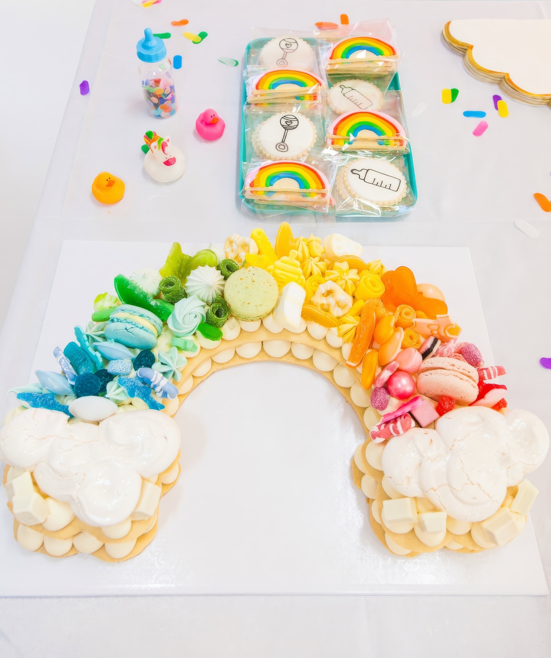 A colorful dessert arrangement on a table features a rainbow-themed cake topped with various candies and macarons. Perfect for a rainbow baby shower, the setup includes decorated cookies with rainbows, airplanes, and baby rattles, a small bottle filled with candy toppings, and colorful confetti pieces scattered around.