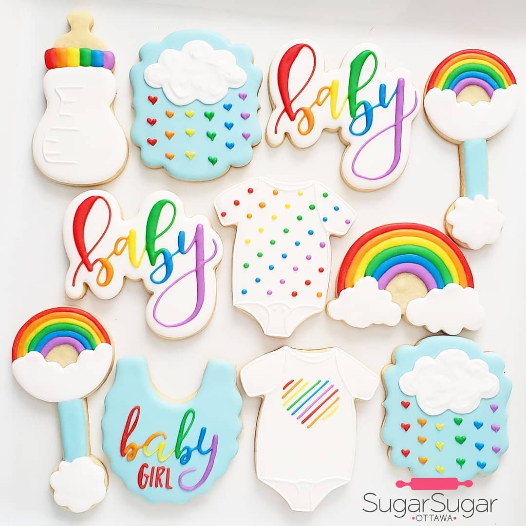 A colorful assortment of sugar cookies decorated for a rainbow baby shower. Shapes include rainbows, clouds, baby bottles, bibs, and onesies. The cookies feature vibrant designs such as hearts, stripes, and cursive "baby" text. A "Sugar Sugar Ottawa" logo is in the corner.