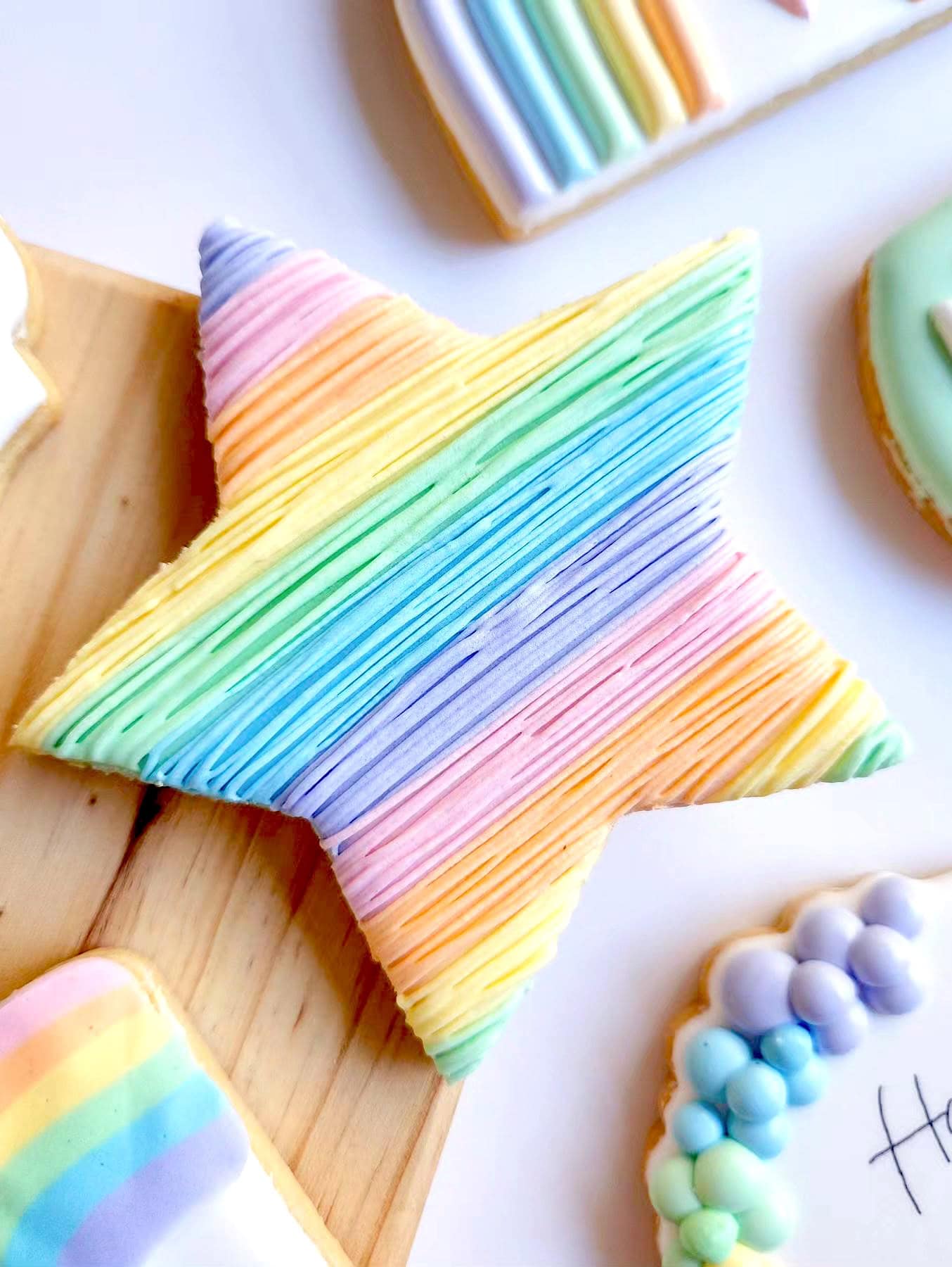 A star-shaped cookie, perfect for a rainbow baby shower, is decorated with icing in vibrant hues arranged in horizontal lines. The cookie rests on a wooden surface alongside other similarly adorned treats featuring pastel colors and delightful rainbow themes.