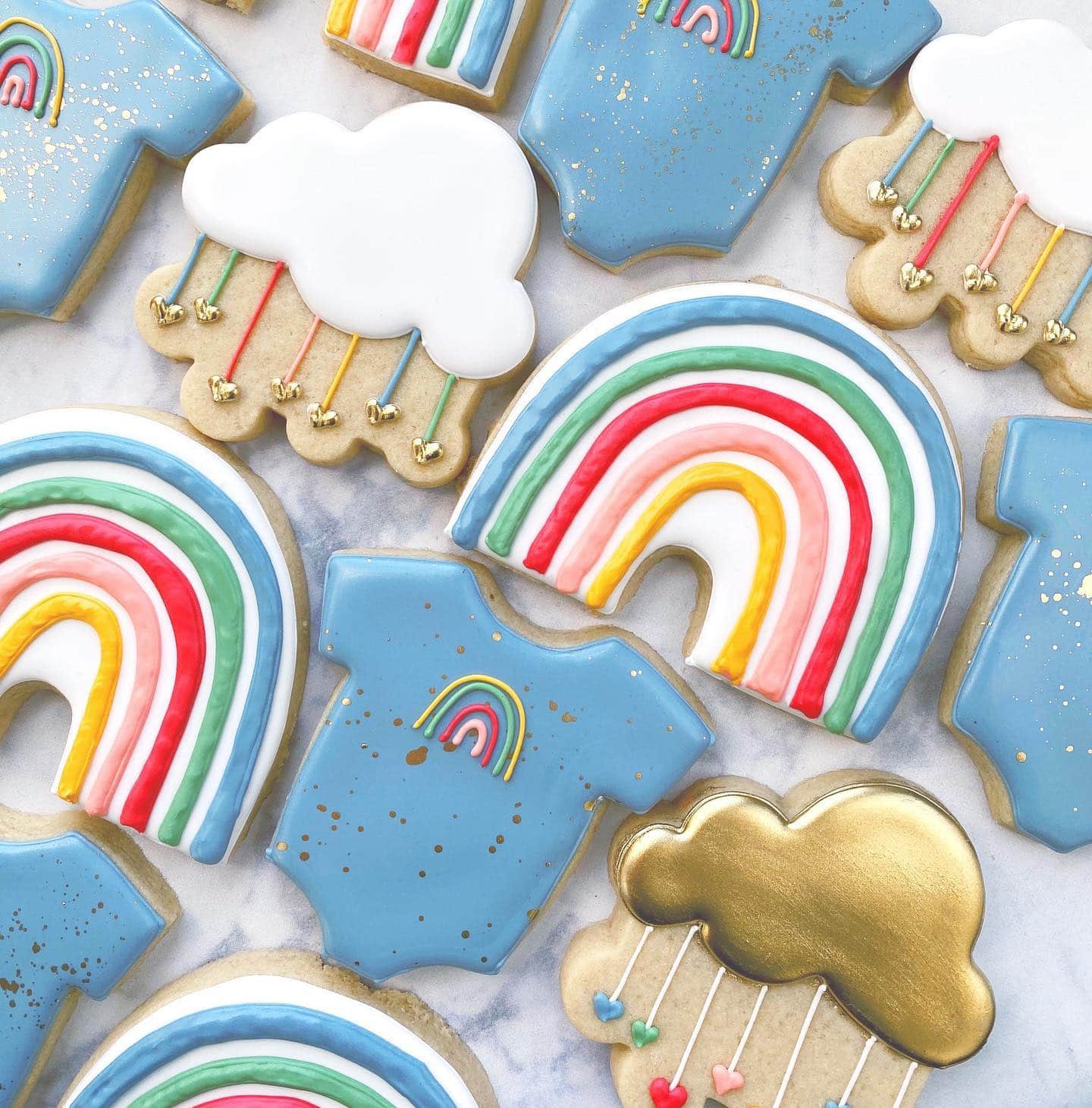 Decorated sugar cookies in various shapes and designs, perfect for a rainbow baby shower. The cookies are shaped like blue onesies, rainbows, white clouds, and golden clouds with multi-colored raindrops. Each cookie is intricately frosted with vibrant pastel colors and delicate details.
