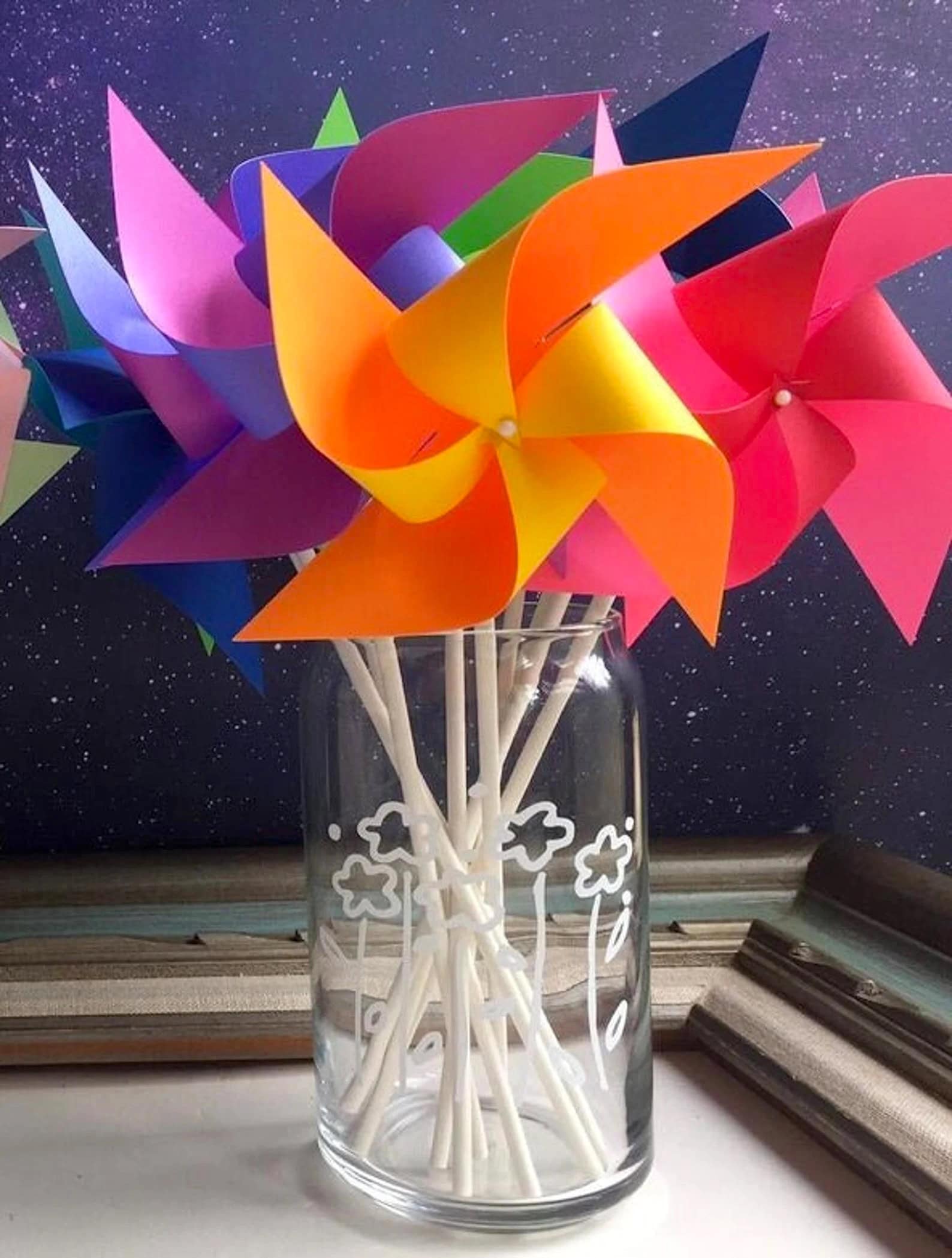 A glass jar filled with colorful paper pinwheels on white sticks is placed on a wooden surface. The pinwheels, perfect for a rainbow baby shower, are in vibrant shades of orange, pink, blue, purple, and yellow. The jar has white decorative drawings and the background features a starry night sky.

