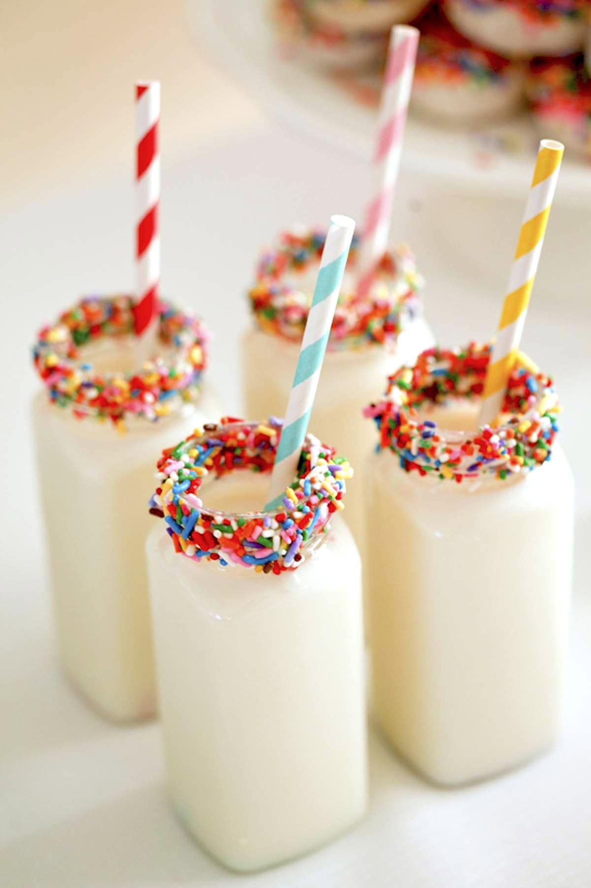 Four glass bottles filled with milk, each topped with a decorative rim covered in colorful sprinkles and a different striped paper straw (red, blue, yellow, and pink), stand on a white surface, creating a festive and playful presentation perfect for a rainbow baby shower.