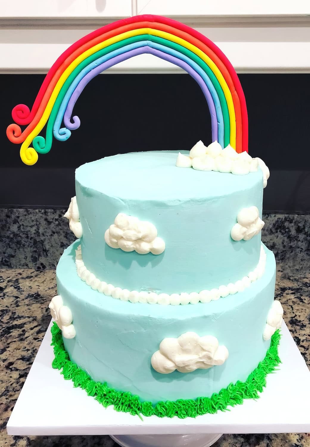 A two-tiered, light blue cake decorated with fluffy white clouds is perfect for a rainbow baby shower. Crowned with a vibrant rainbow made from fondant and surrounded by green frosting grass at the base, it adds a touch of magic as the rainbow arches high above, curving gracefully.