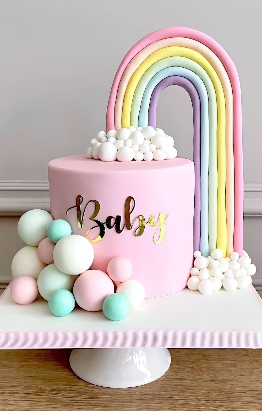 A pastel-themed cake perfect for a rainbow baby shower, with "Baby" written in gold on the side. The cake features a rainbow made of fondant extending from the top to the side, surrounded by white clouds. Pastel-colored spheres decorate the base of the cake, which is set on a white cake stand.