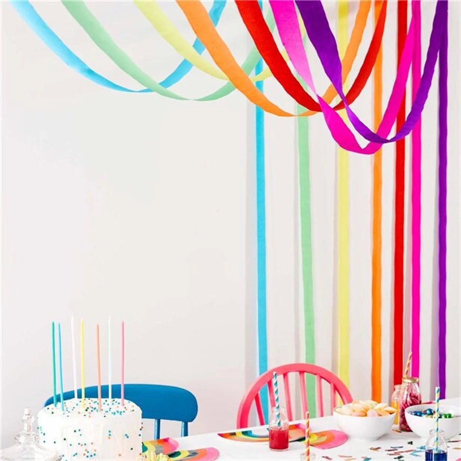 A vibrant rainbow baby shower setup with rainbow streamers hanging from the ceiling. A white cake with pastel candles sits on the table, surrounded by bowls of snacks and drinks with striped straws. Blue and pink chairs are at the table, which has delightful rainbow-themed decor.
