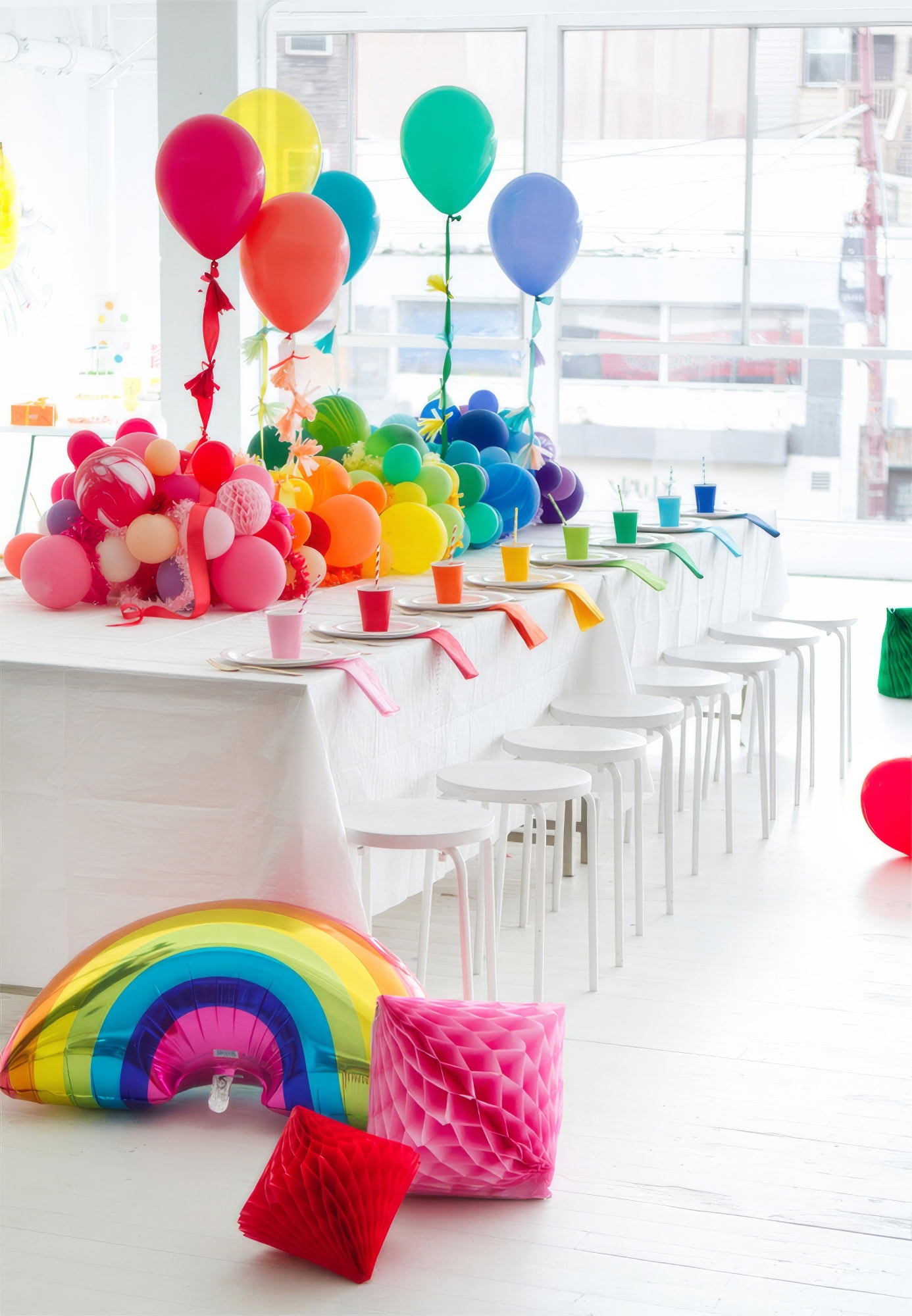 A brightly decorated party room with a long table covered in a white cloth awaits guests at the rainbow baby shower. The table features colorful balloons and cups arranged in a rainbow pattern. Chairs are lined up along the table, and a large rainbow balloon and colorful honeycomb decorations adorn the floor.