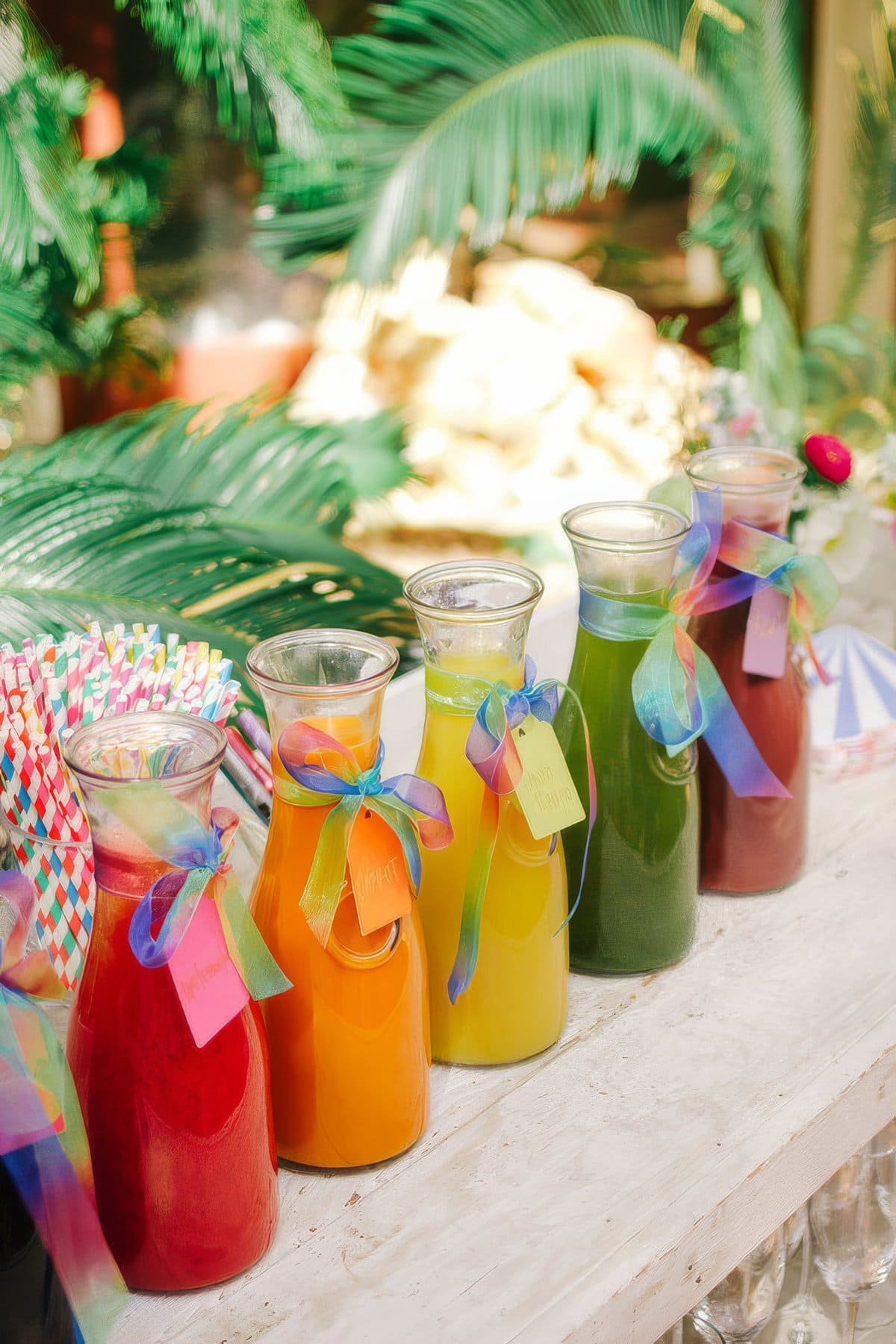 Five glass carafes containing different brightly colored juices are lined up on a white wooden surface. Each carafe has a ribbon and a label attached. The background, perfect for a rainbow baby shower, is decorated with large green leaves and vibrant multicolored straws.