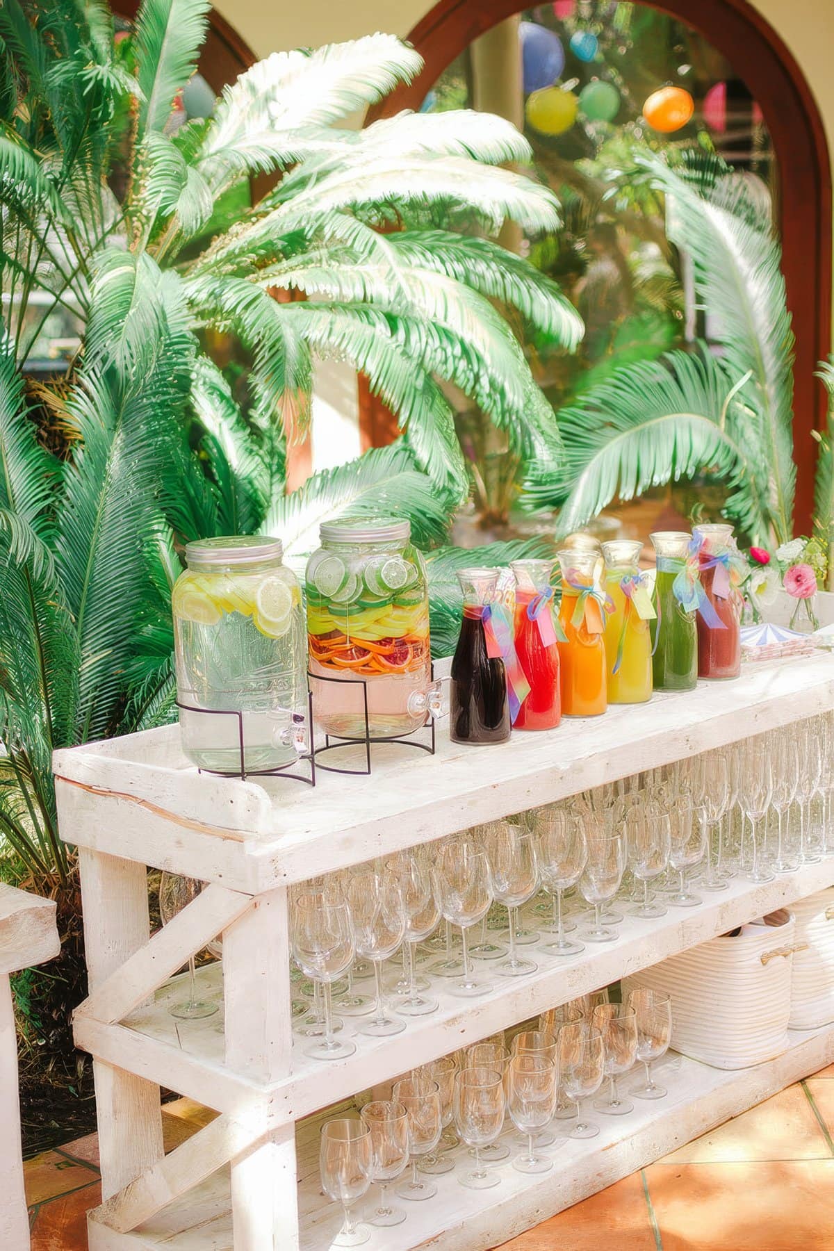 A wooden stand holds jars and pitchers of colorful beverages, including water with lemon and various fruit juices. Below, rows of empty wine glasses are ready to be filled. Lush green plants and vibrant decorations set the scene for a festive rainbow baby shower.