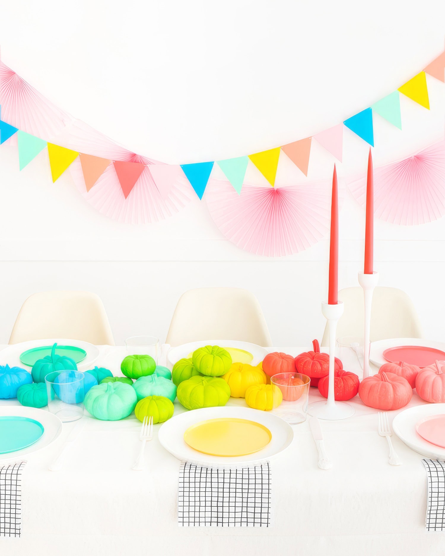 A brightly decorated table set for a rainbow baby shower features colorful pumpkins in green, blue, yellow, and pink. Two tall red candles in white holders are centered on the table. Background decorations include multicolored triangular pennant banners and pink fans.