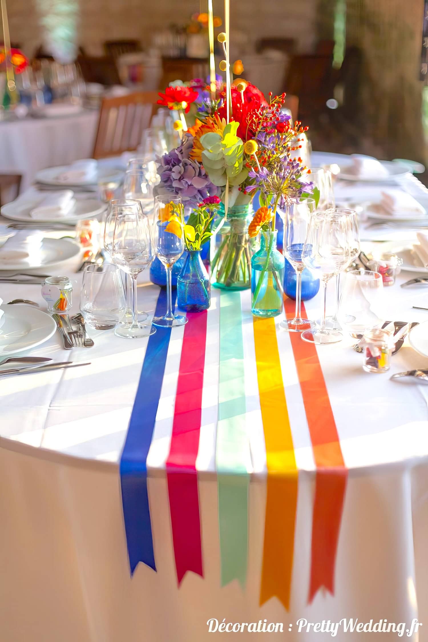 A decorated wedding table showcases rainbow baby shower vibes with blue, pink, red, orange, yellow, and green ribbons extending from the centerpiece. Vibrant flowers in glass vases complement the elegant dinnerware, wine glasses, and cutlery neatly arranged around the table. Text in the bottom right corner reads "Décoration : PrettyWedding.fr.