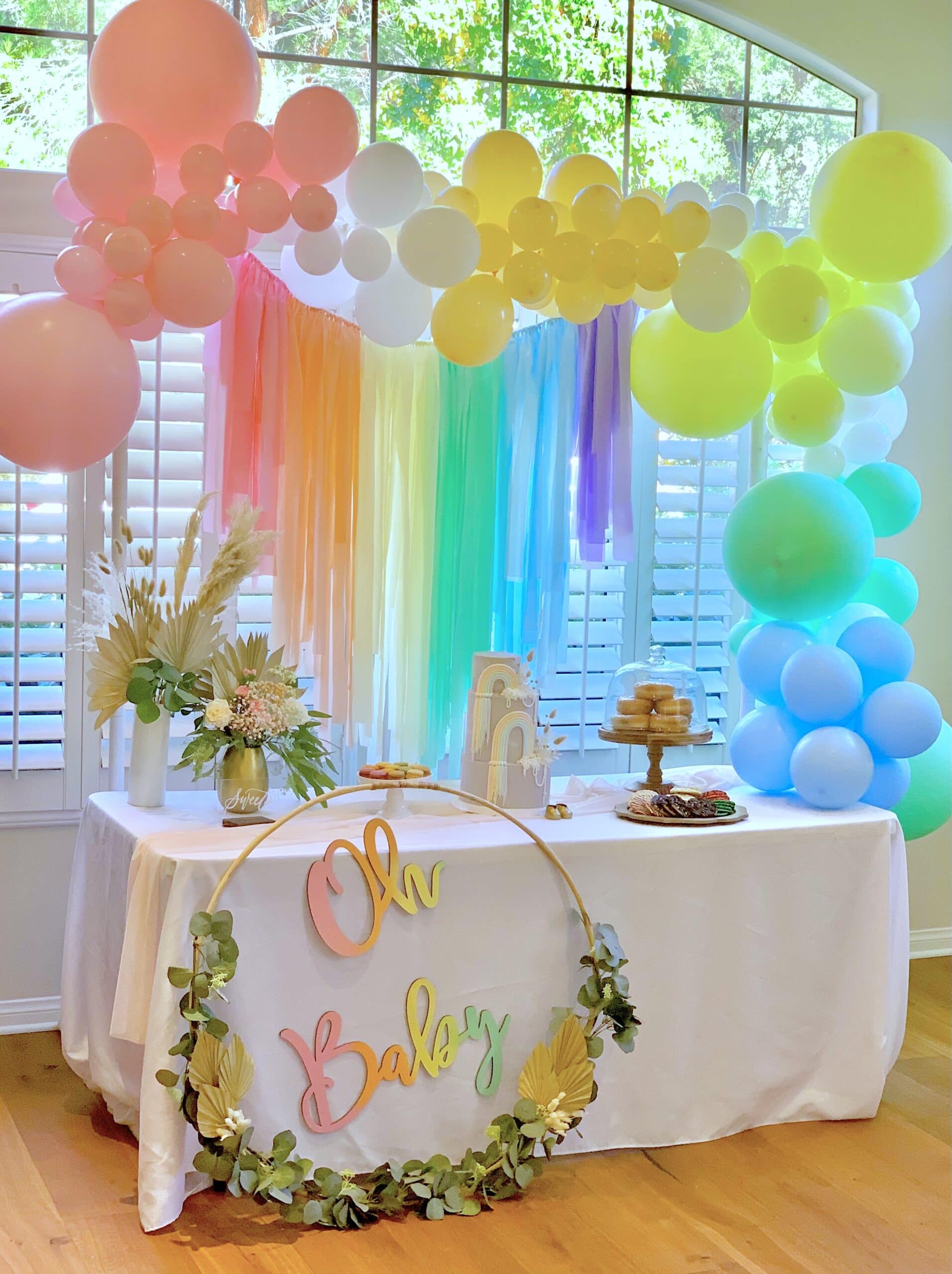 A rainbow baby shower setup features a table covered in a white cloth, adorned with a floral centerpiece, cakes, and treats. Above, a rainbow backdrop and pastel balloons decorate the area. A circular sign with flowers reads "Oh Baby." Large windows with shutters provide the backdrop.