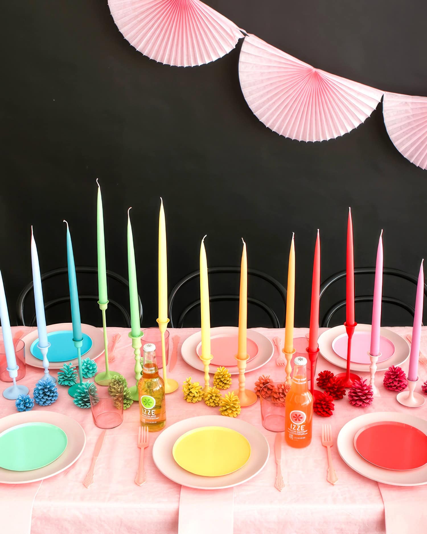 A festive table setting with colorful decor is perfect for a rainbow baby shower. Tall, pastel-colored taper candles in gradient hues adorn the center. Plates and utensils in matching soft tones are arranged neatly. Two bottled drinks are also visible. Pink paper fans hang as decoration in the background.