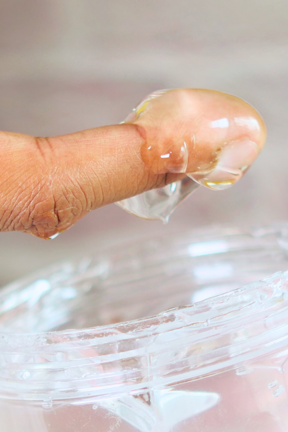 A close-up of a person's finger holding a dollop of clear, diy hair gel over an open container. The gel is thick and shiny, stretching slightly between the finger and the container. The background is blurred and neutral in color.