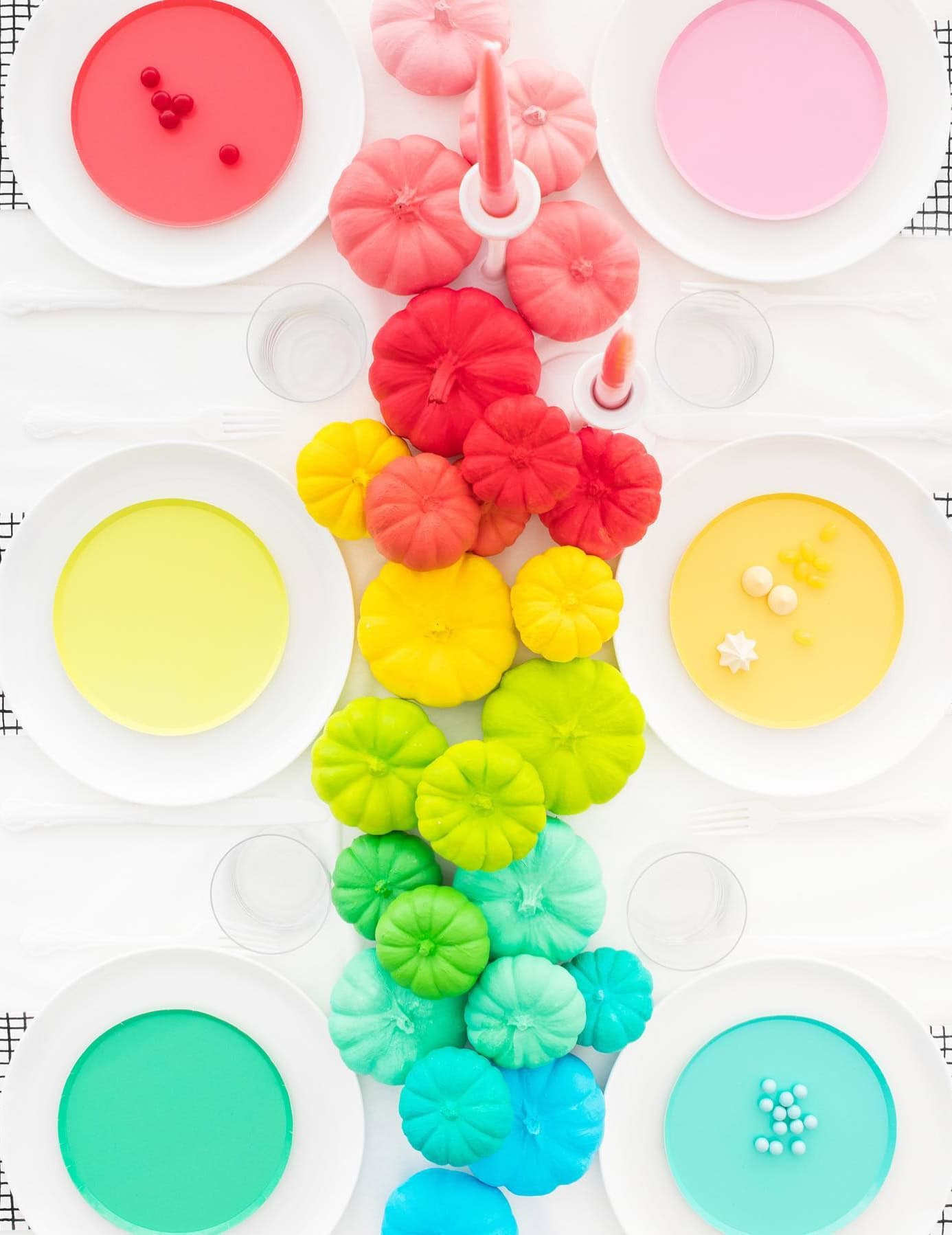 A brightly colored table setting for a rainbow baby shower features a white tablecloth and plates with pastel-colored settings. Miniature pumpkins painted in a gradient of rainbow colors are arranged down the center. The decorations include candles and small confectioneries, adding whimsical charm.