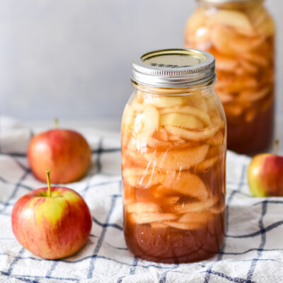 A jar filled with preserved apple slices in a syrupy liquid sits on a white and blue checkered cloth, ready to be used for easy apple pie filling. Surrounding the jar are three fresh apples, while another similar jar is blurred in the background.