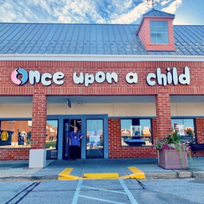 A store with a brick facade called Once Upon a Child specializes in used clothing. A person stands in the doorway, waving. Large windows display various items, and flowers bloom in planters by the entrance. The sky is blue with a few clouds.