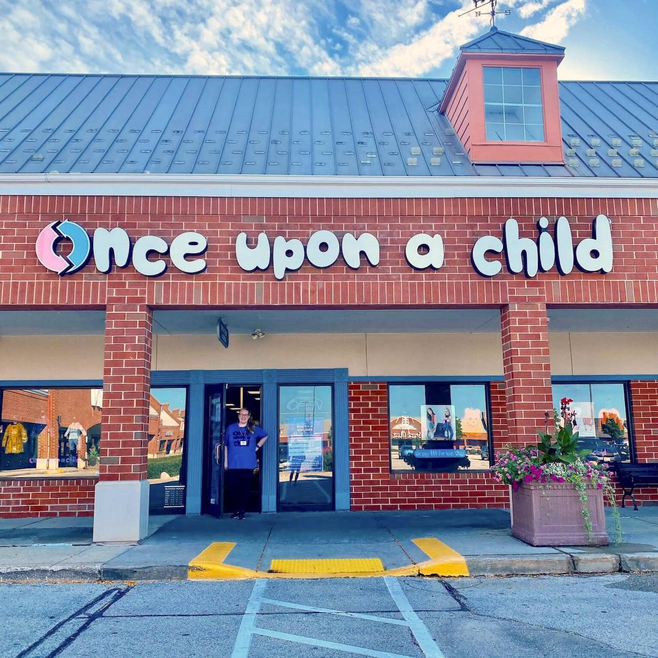 A store with a brick facade called Once Upon a Child specializes in used clothing. A person stands in the doorway, waving. Large windows display various items, and flowers bloom in planters by the entrance. The sky is blue with a few clouds.