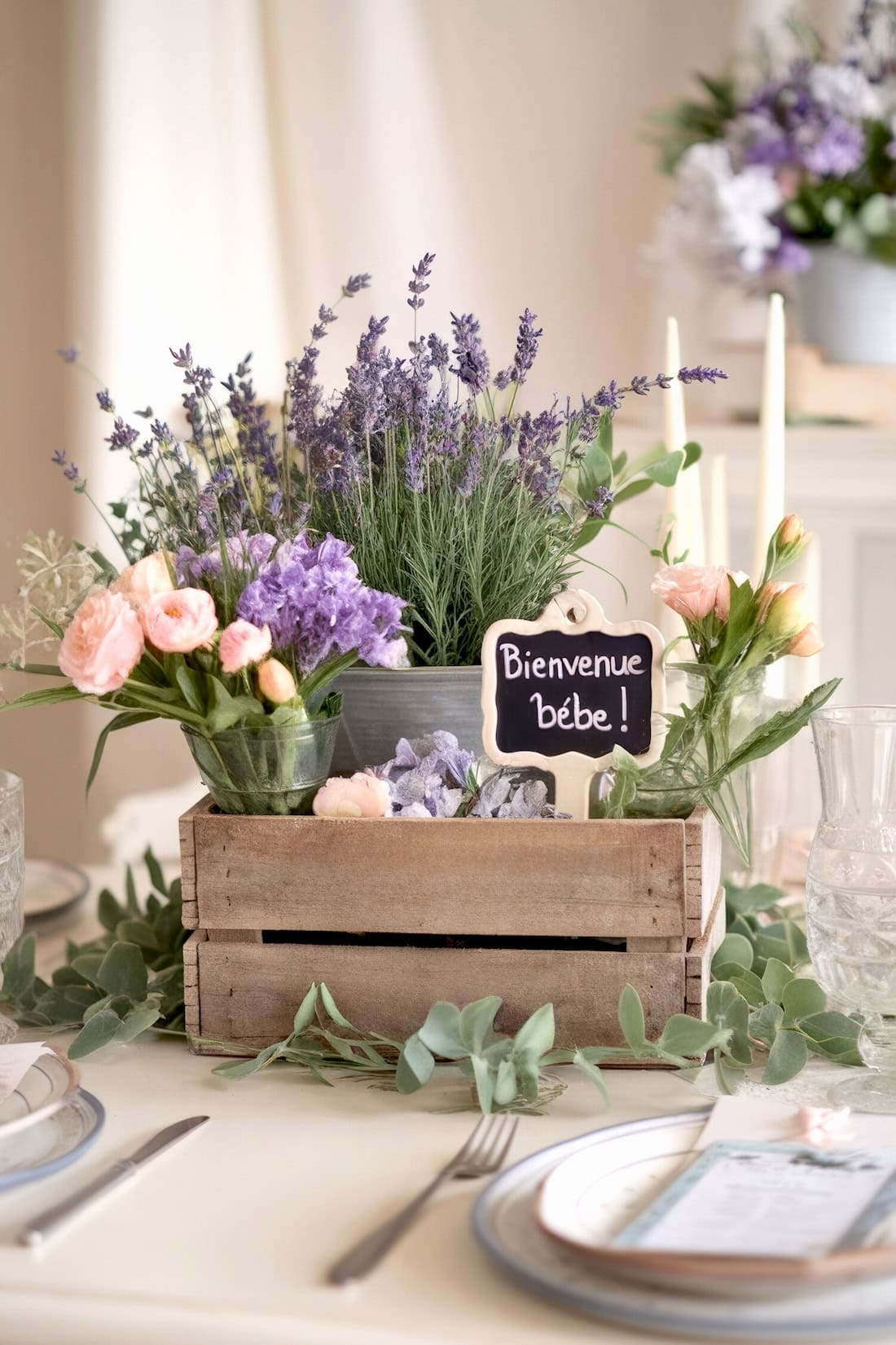 A rustic wooden crate on a table holds lavender and pink baby shower centerpiece flowers, with a small chalkboard sign reading "Bienvenue bébé!" Plates with menus and green leaves adorn the table, creating an elegant and welcoming setting.