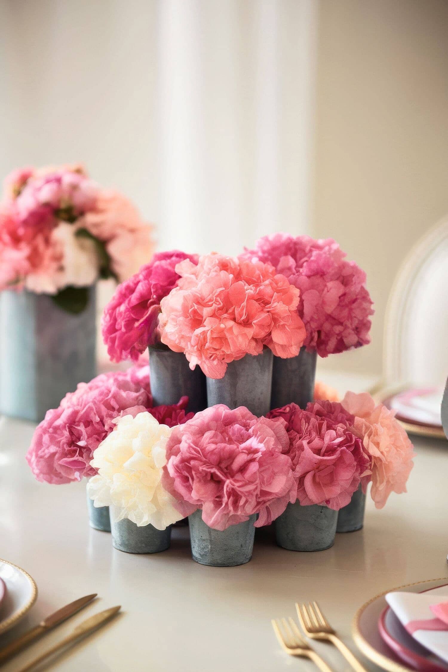 A baby shower floral arrangement centerpiece featuring pink, white, and peach carnations in small gray vases graces a dining table set with gold cutlery and elegant pink and white tableware.