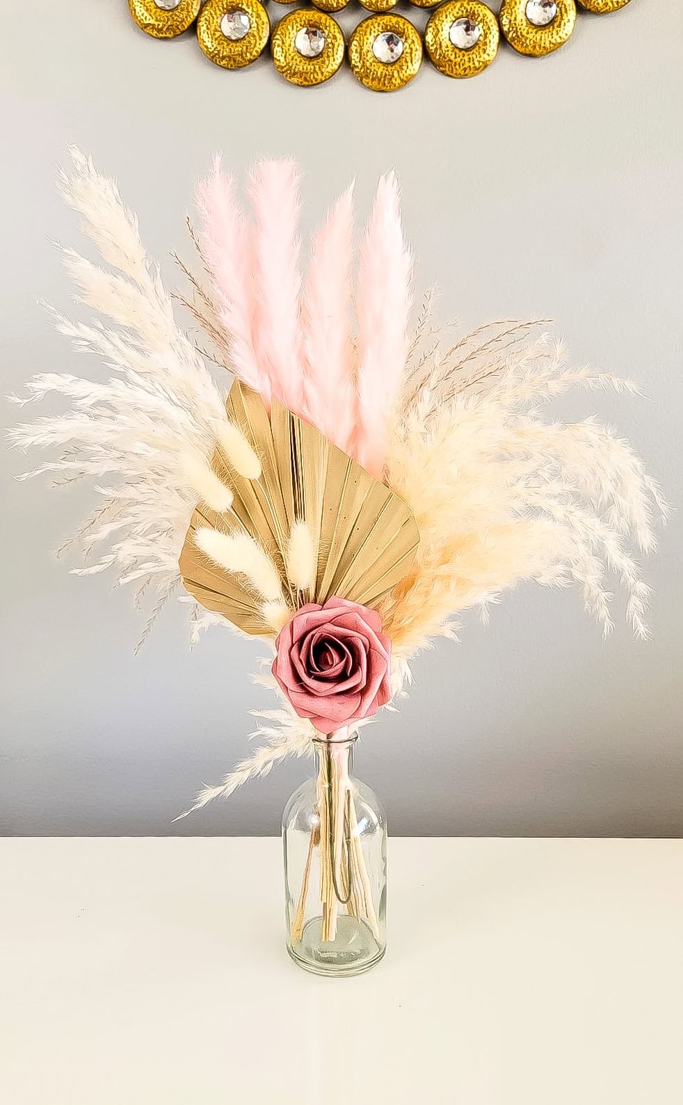 A delightful baby shower centerpiece idea features a decorative arrangement in a glass vase with a pink rose, beige and pink pampas grass, and a metallic gold fan-shaped leaf set against a light gray background.
