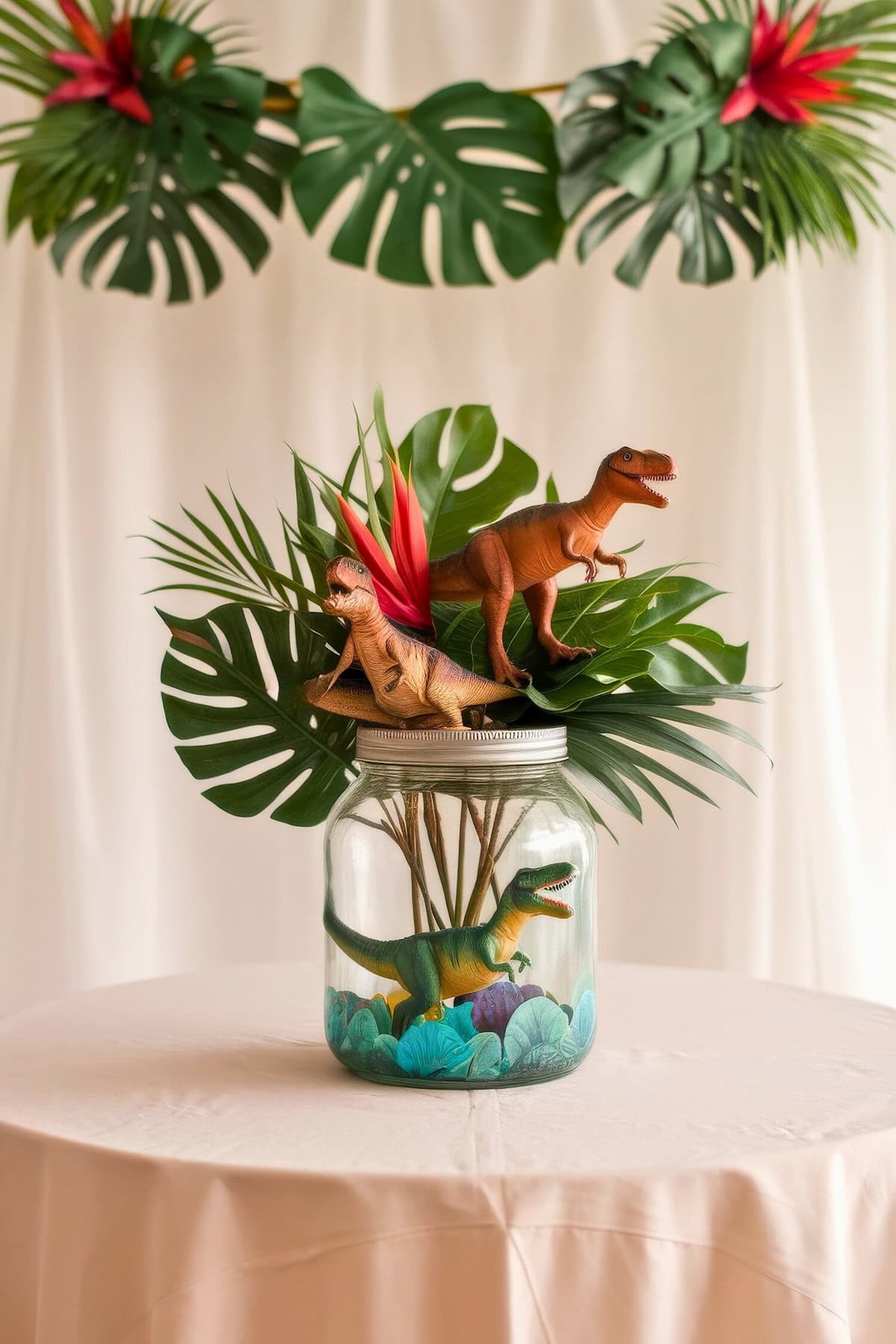 A decorative jar on a table, perfect for baby shower centerpieces, contains tropical leaves and toy dinosaurs, adorned with red and pink flowers. Above, a garland of green leaves and red flowers hangs playfully, enhancing the nature-themed display.