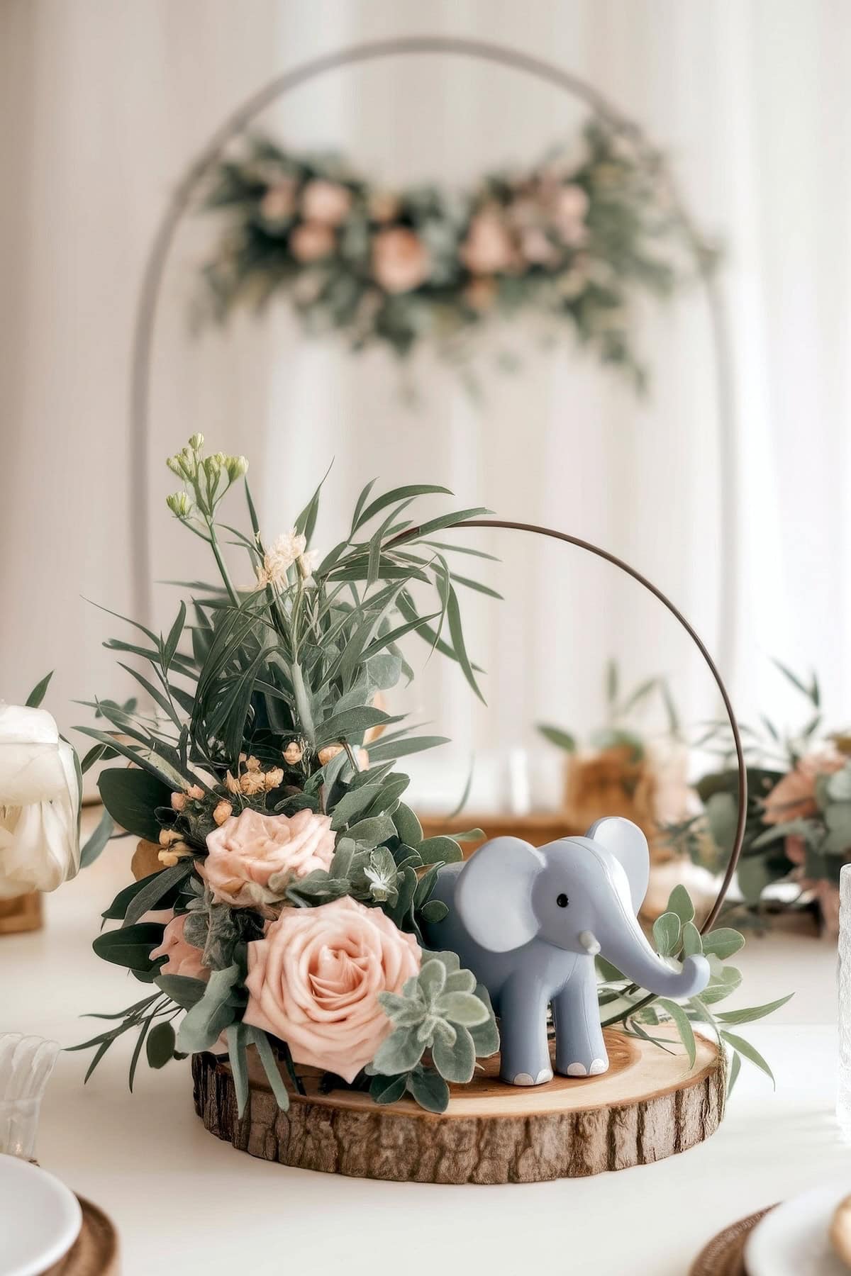A whimsical baby shower centerpiece featuring a small blue elephant figurine surrounded by lush greenery and pink roses, set on a round wooden base. A floral arrangement in the background completes the elegant decor.