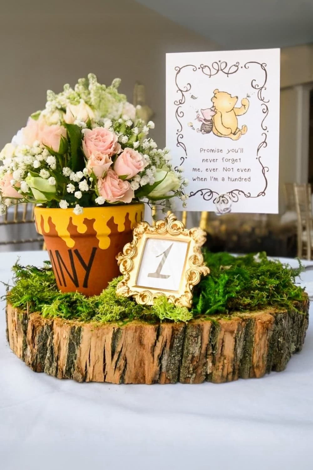 A charming baby shower centerpiece featuring pastel roses and white flowers in a pot labeled "Hunny." A framed number "1" rests on moss, and beside it, a card with a bear reads, "Promise you'll never forget me, ever. Not even when I'm a hundred.
