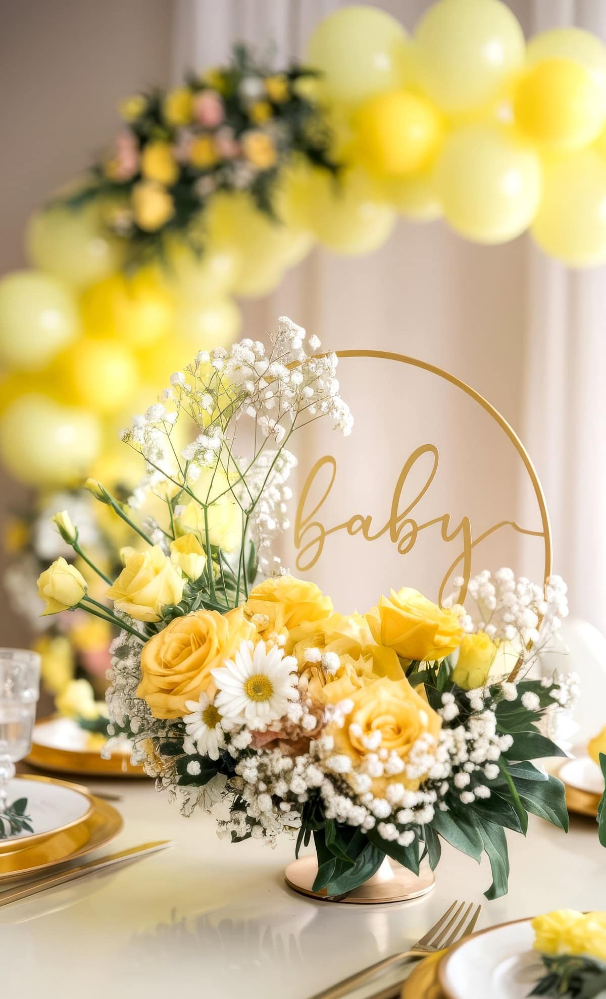 A floral centerpiece featuring yellow roses, daisies, and baby's breath artfully arranged around a circular "baby" sign serves as the perfect baby shower centerpiece. In the background, a cheerful yellow balloon arch enhances the festive atmosphere of this delightful celebration.