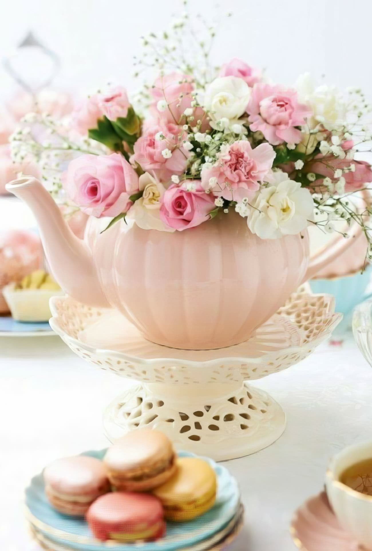 A pastel pink teapot is elegantly arranged with pink and white roses, baby's breath, and carnations, perfect for baby shower centerpieces. It sits on a decorative white stand, while colorful macarons are artfully displayed on a blue plate in the foreground.