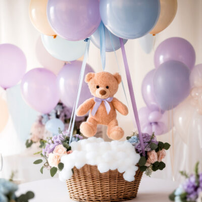 A teddy bear sits in a wicker basket surrounded by purple and pink flowers, with pastel balloons lifting the basket. Perfect as baby shower centerpieces, the setting is festive, with more balloons in the background creating a whimsical atmosphere.