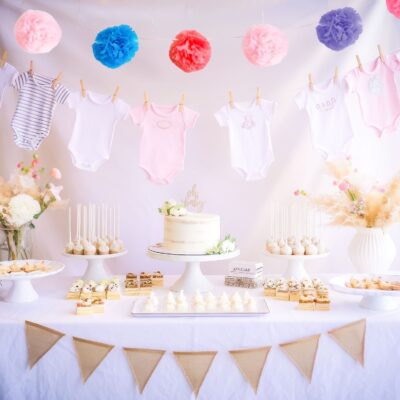 A baby shower table brimming with delightful ideas showcases a cake, cake pops, and pastries. Overhead, colorful pom-poms and charming baby onesies hang on a line. The table radiates joy with its floral accents and a cheerful triangular banner.