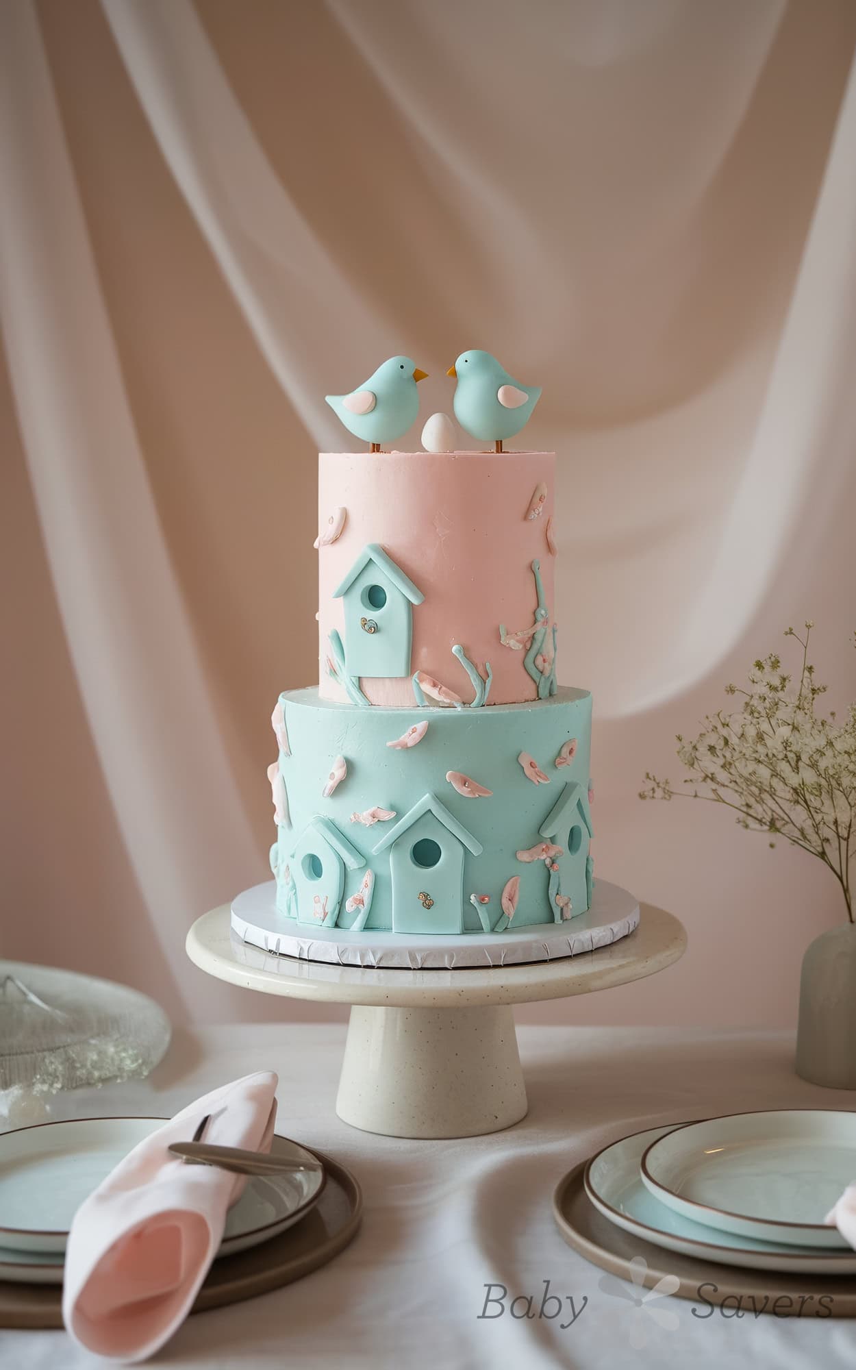 A two-tiered cake on a stand, with pink and blue fondant, offers delightful bird baby shower ideas. The bottom tier showcases a charming birdhouse design with tiny birds, while the top features two blue fondant birds. The table is elegantly set with plates, napkins, and a small vase of flowers.