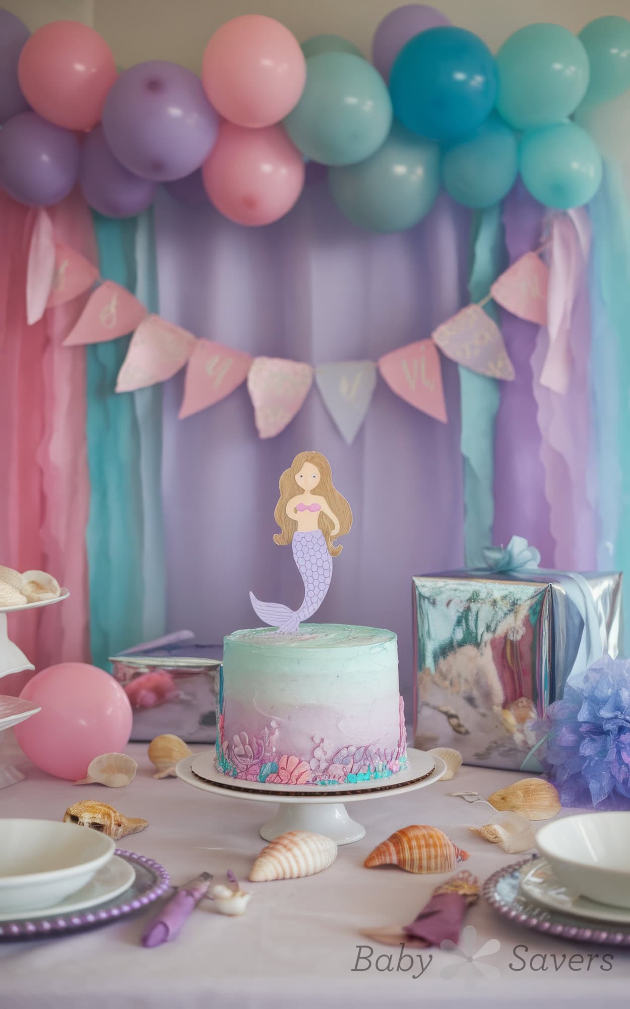 A beautifully decorated mermaid-themed party table, perfect for baby shower ideas, features a pastel gradient cake topped with a mermaid and surrounded by seashells. The backdrop consists of purple, pink, and blue balloons and streamers, with a banner and gift box in the background.
