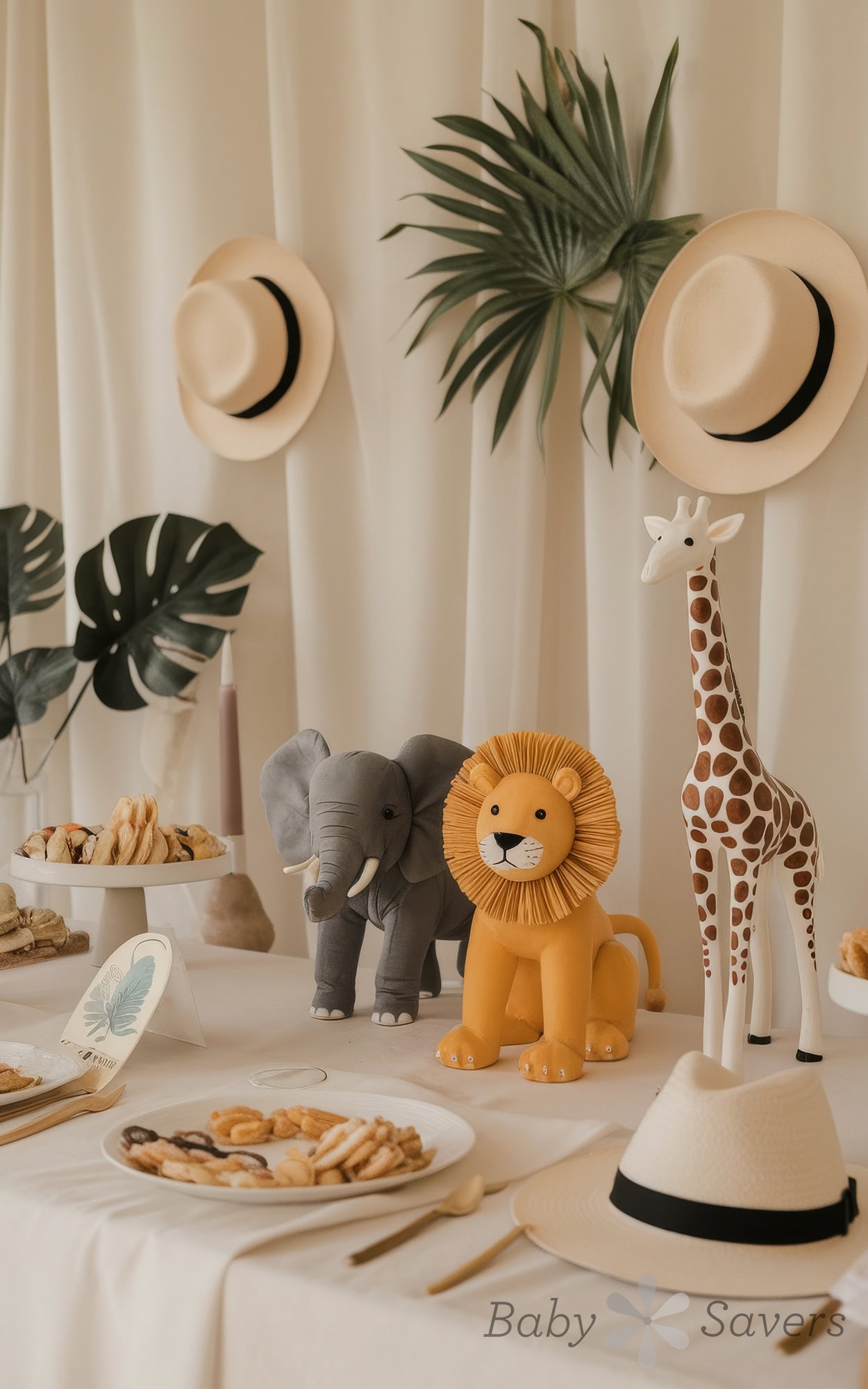 A safari-themed baby shower theme decoration features plush animals like an elephant, lion, and giraffe on a white table. Straw hats hang in the background with large green leaves, while plates of cookies and pastries add to the delightful spread.