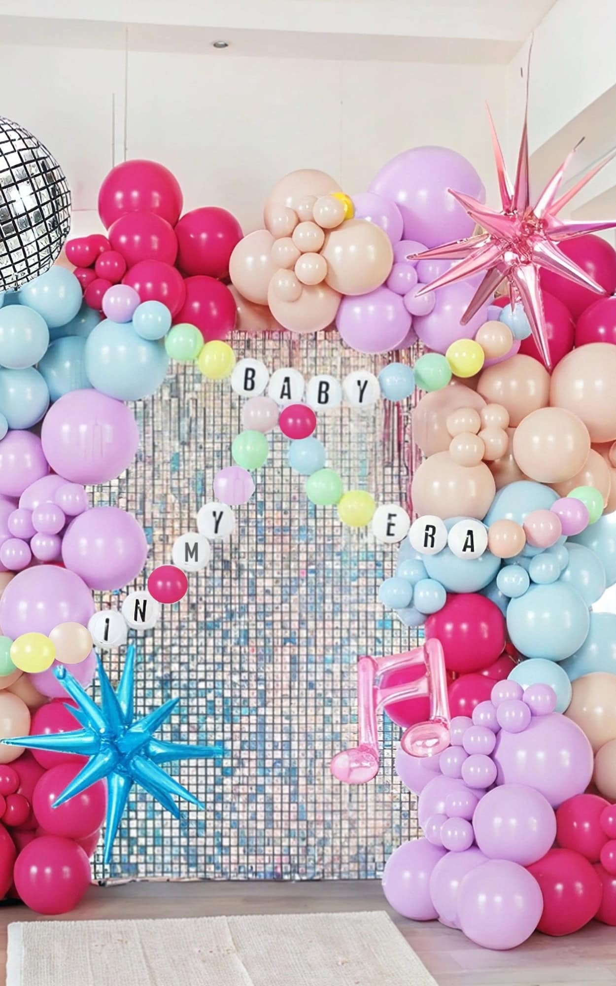 A colorful balloon arch featuring pink, red, beige, blue, and purple balloons surrounds a beaded curtain for a taylor swift baby shower. Metallic star shapes and music notes add flair—perfect for baby shower ideas. A banner reads "IN MY BABY ERA," with a reflective disco ball hanging above.