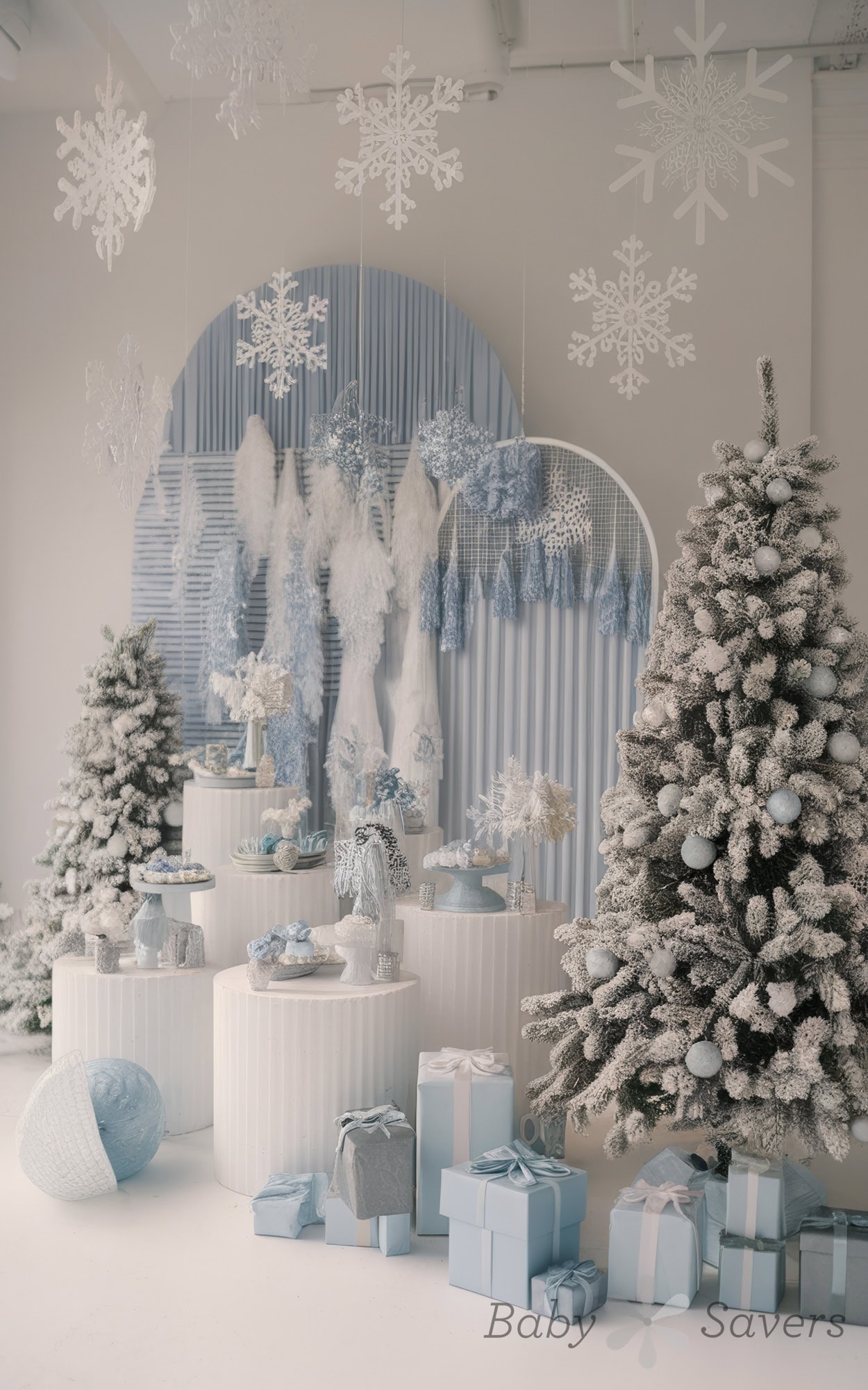 A winter-themed display, perfect for a winter wonderland baby shower idea, features snow-dusted Christmas trees, white snowflakes hanging above, and a collection of blue and silver wrapped gifts. The backdrop includes white and blue decorations, creating a festive yet elegant atmosphere.