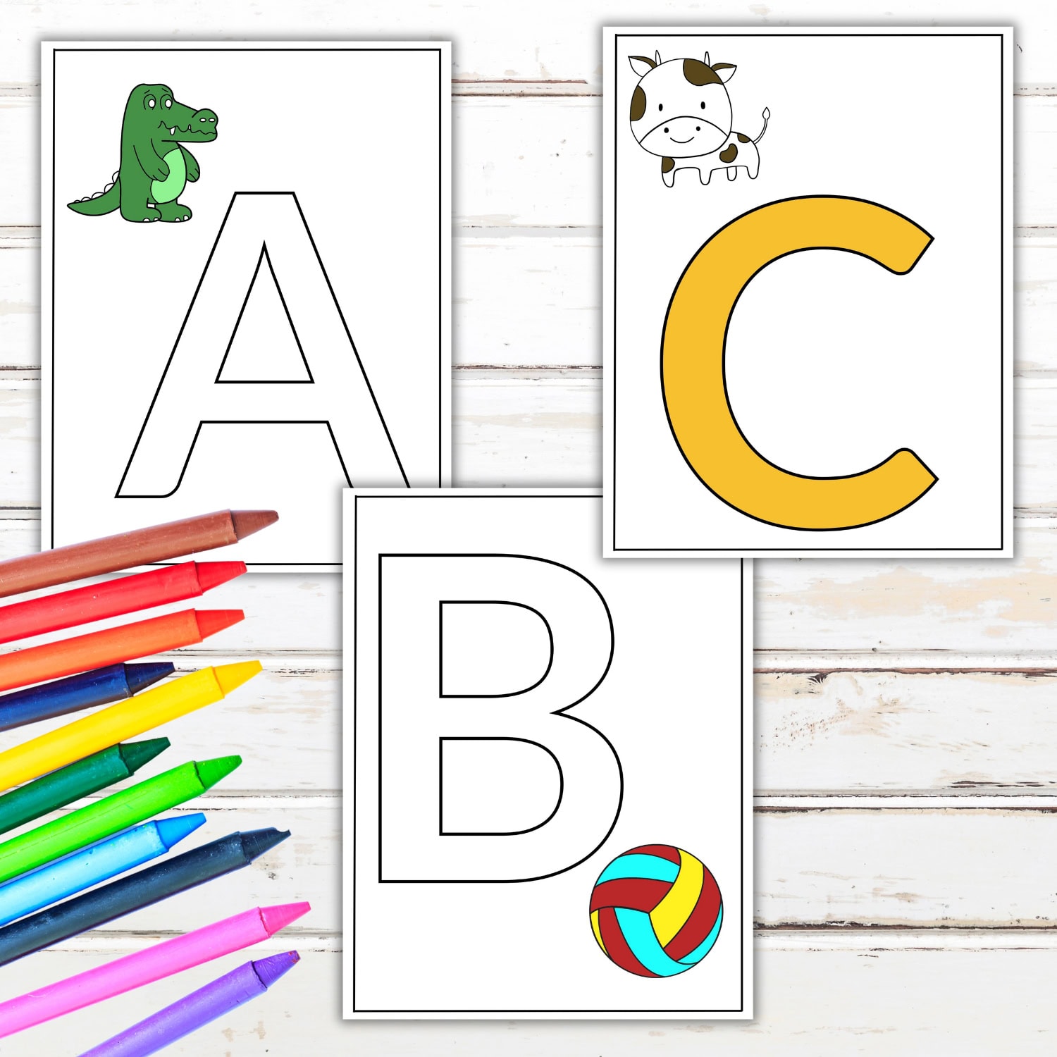 Three cards with the letters A, B, and C. The A card features a green alligator, the B card displays a colorful beach ball, and the C card has a cartoon cow. A set of colorful crayons awaits beside these playful alphabet coloring pages on a wooden surface.
