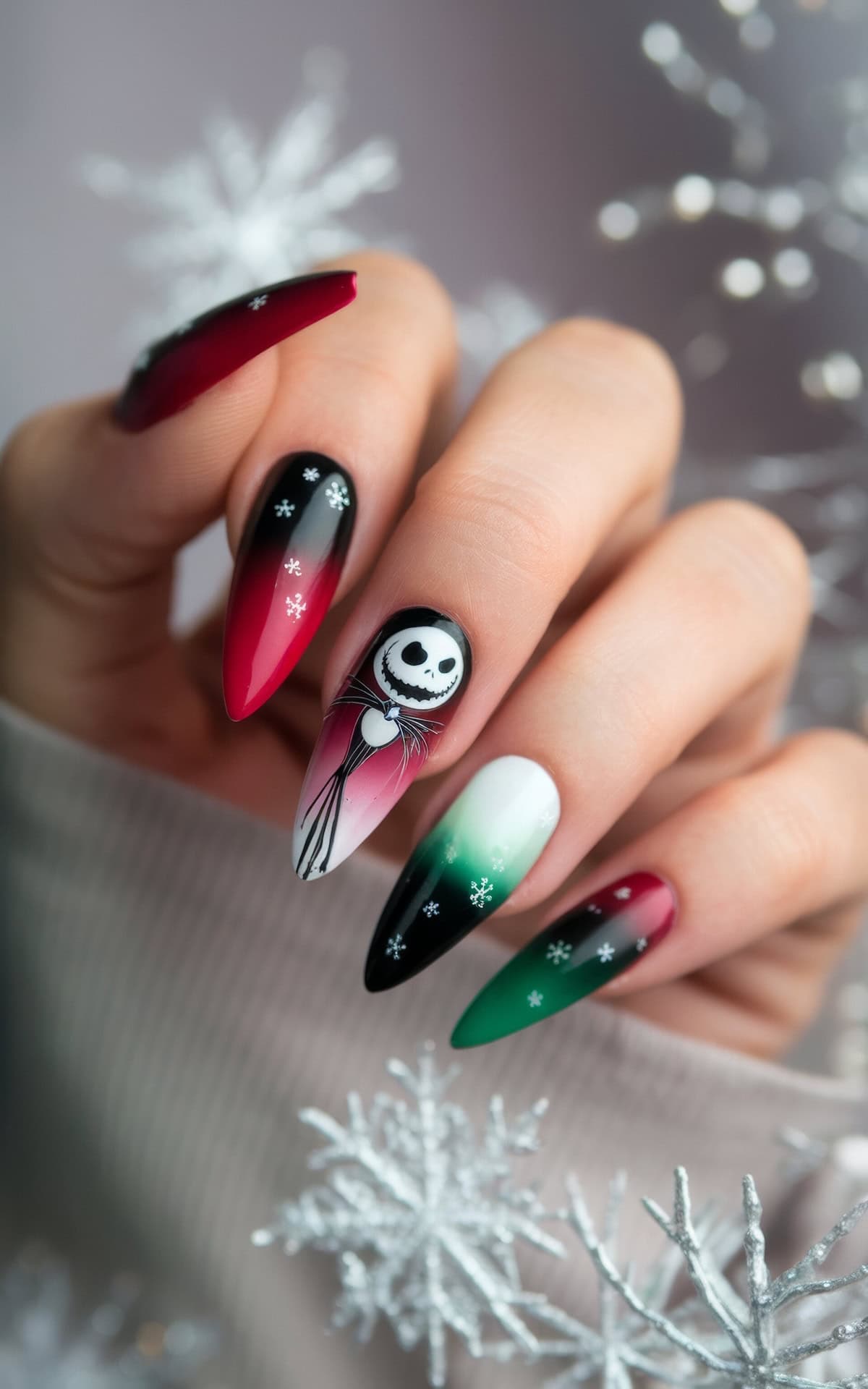 A hand with long nails featuring festive Disney Christmas nail art. The nails showcase a mix of red, green, black, and white colors, with designs including snowflakes and a Jack Skellington from The Nightmare Before Christmas face. The background has a soft focus with snowflake decorations.