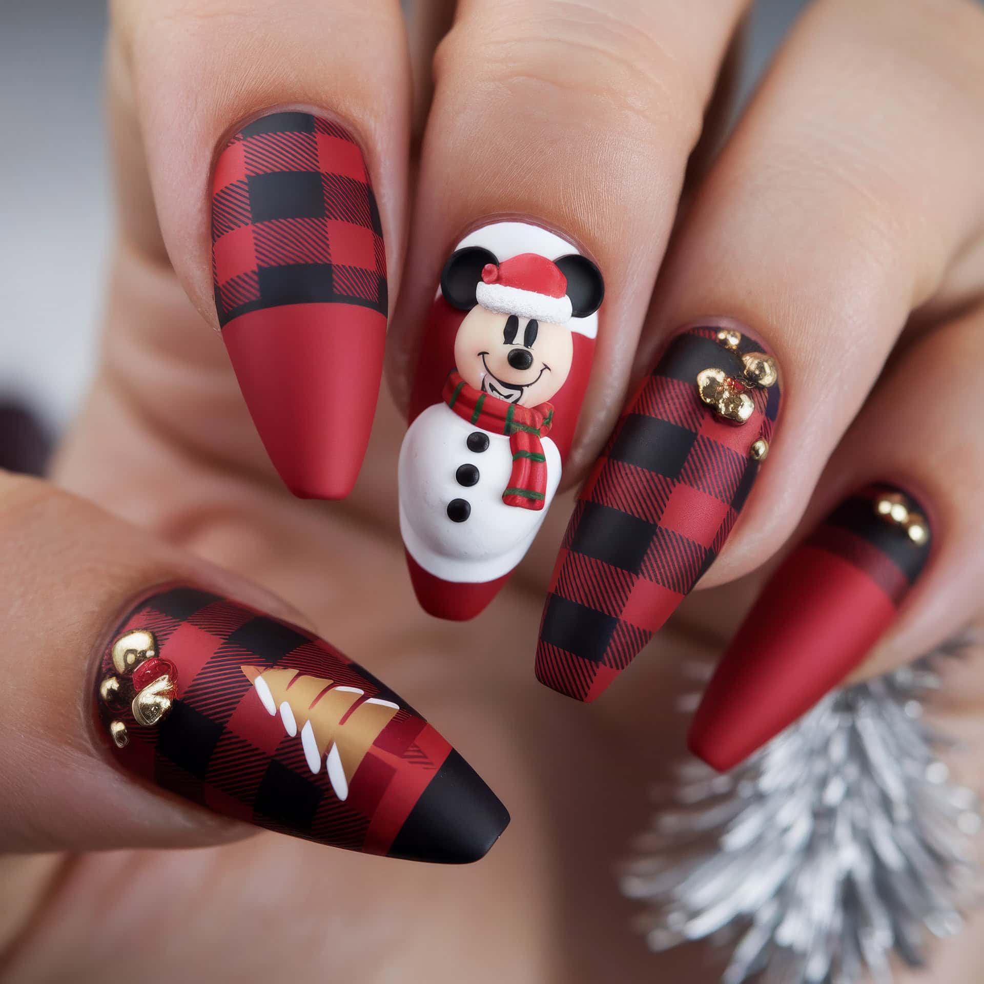 Close-up of festive Disney Christmas nails showcasing red and black plaid patterns, a mouse ears snowman design with a Mickey mouse face, and gold decorations resembling a Christmas tree. The nails have a matte finish and a pointed shape.