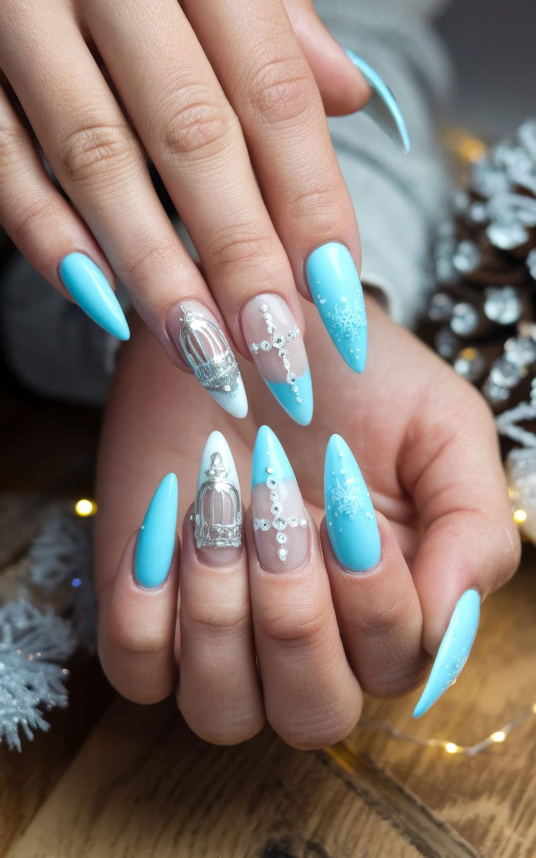 Close-up of hands with long, pointed nails painted in light blue and white. Nails showcase intricate Disney Cinderella Christmas-themed designs, including snowflakes, pearls, and decorative silver accents, all set against a wooden background with festive elements.