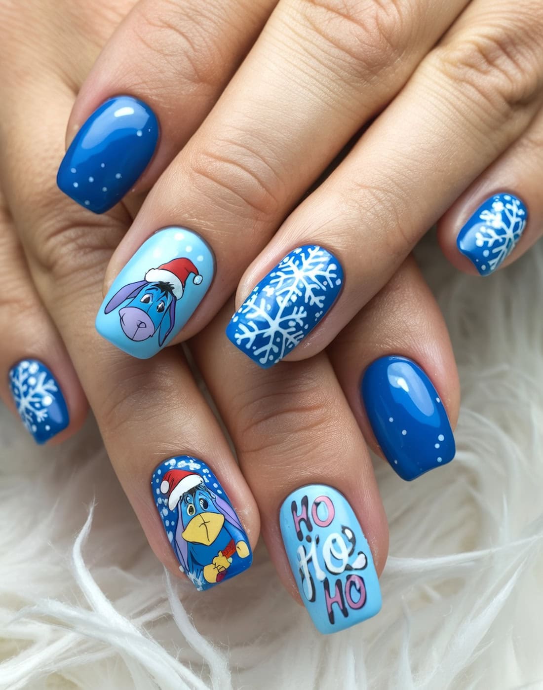Close-up of festive Disney Christmas nails featuring blue and white snowflakes, a cartoon eeyore from Winnie the Pooh in a Santa hat, and "Ho Ho Ho" text. The glossy finish enhances each design against the soft white fabric background.