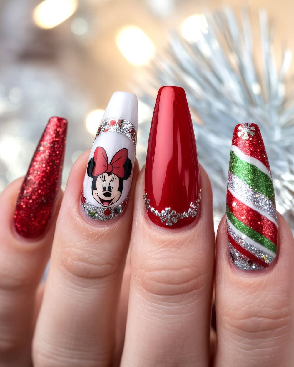 A hand with festive Disney Christmas nail art: one nail sparkles with red glitter, another features a Minnie mouse with a bow, alongside a glossy red nail adorned with snowflakes and a striped design of red, green, and silver glitter reminiscent of a candy cane. The background is softly blurred.
